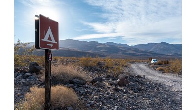 Symbol for tent on a small brown sign on a post stands left of a dirt road. White clouds dot sky.