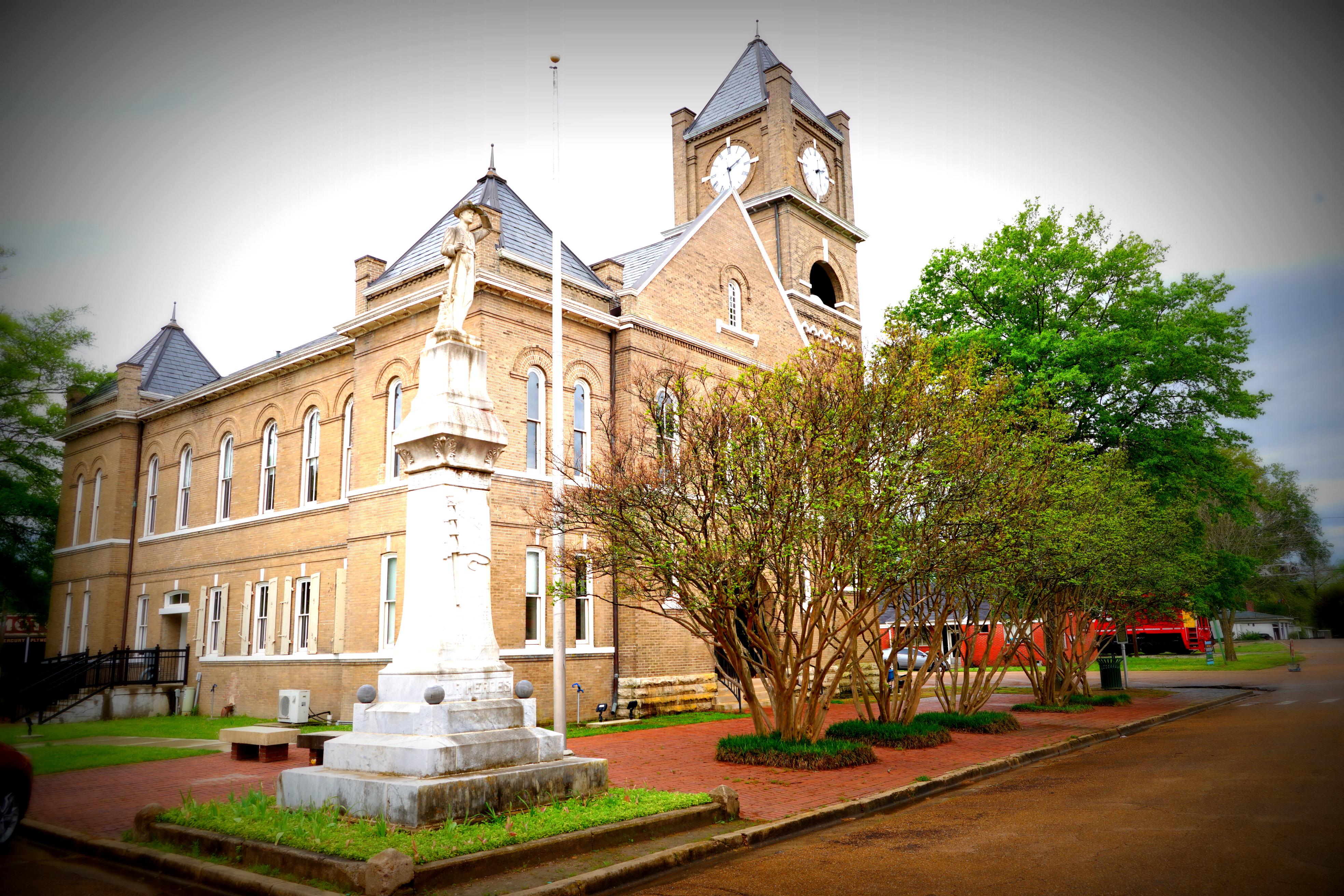Side view of a two-story brick courthouse with a clock tower. A statue stands in front.