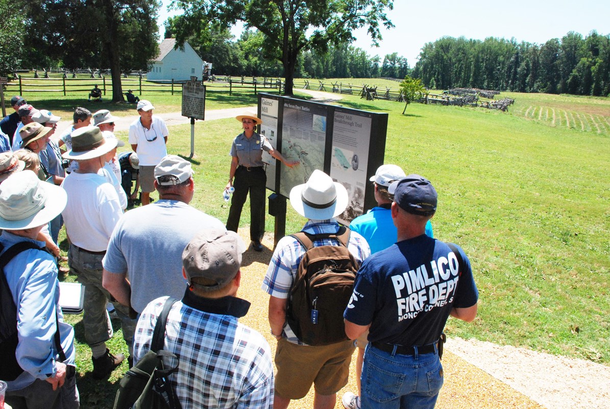 A uniformed park ranger gestures to an upright sign in front of a crowd of visitors.