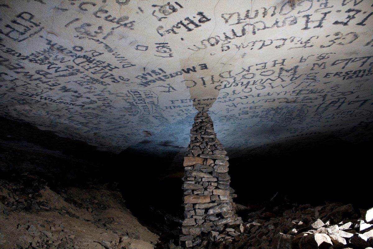 A large stacked stone pillar reaches the flat ceiling containing signatures in a cave passage. 