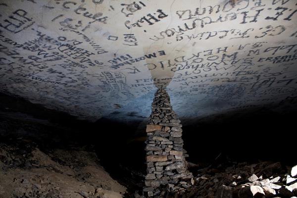 A large stacked stone pillar reaches the flat ceiling containing signatures in a cave passage.