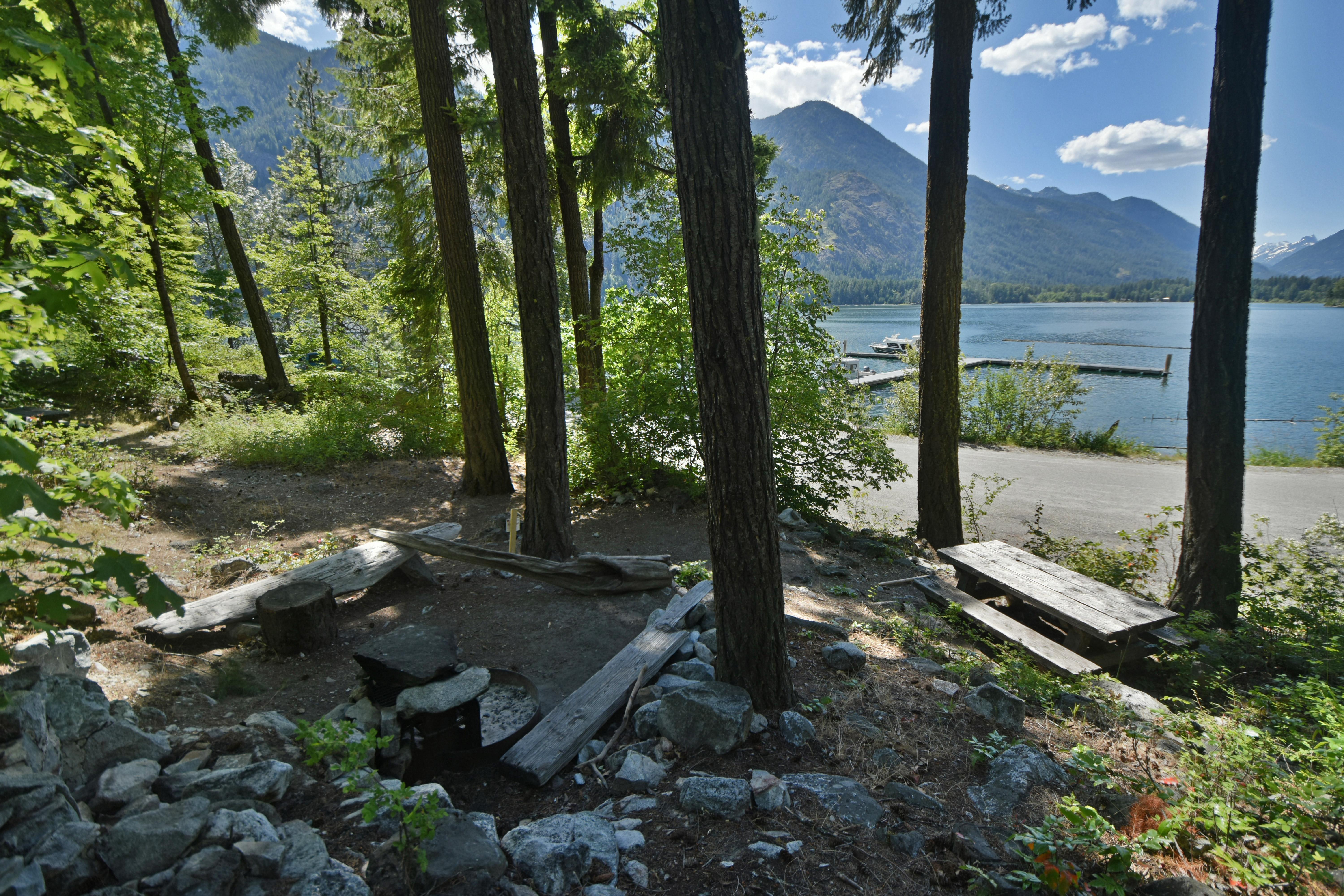 A wooden picnic table at a campsite overlooks a paved road, large lake, and distant mountains.