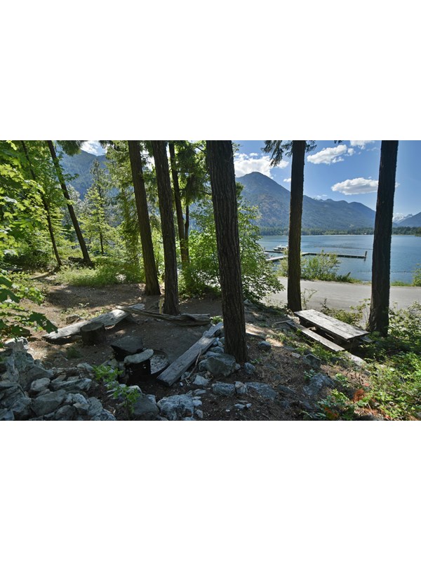 A wooden picnic table at a campsite overlooks a paved road, large lake, and distant mountains.