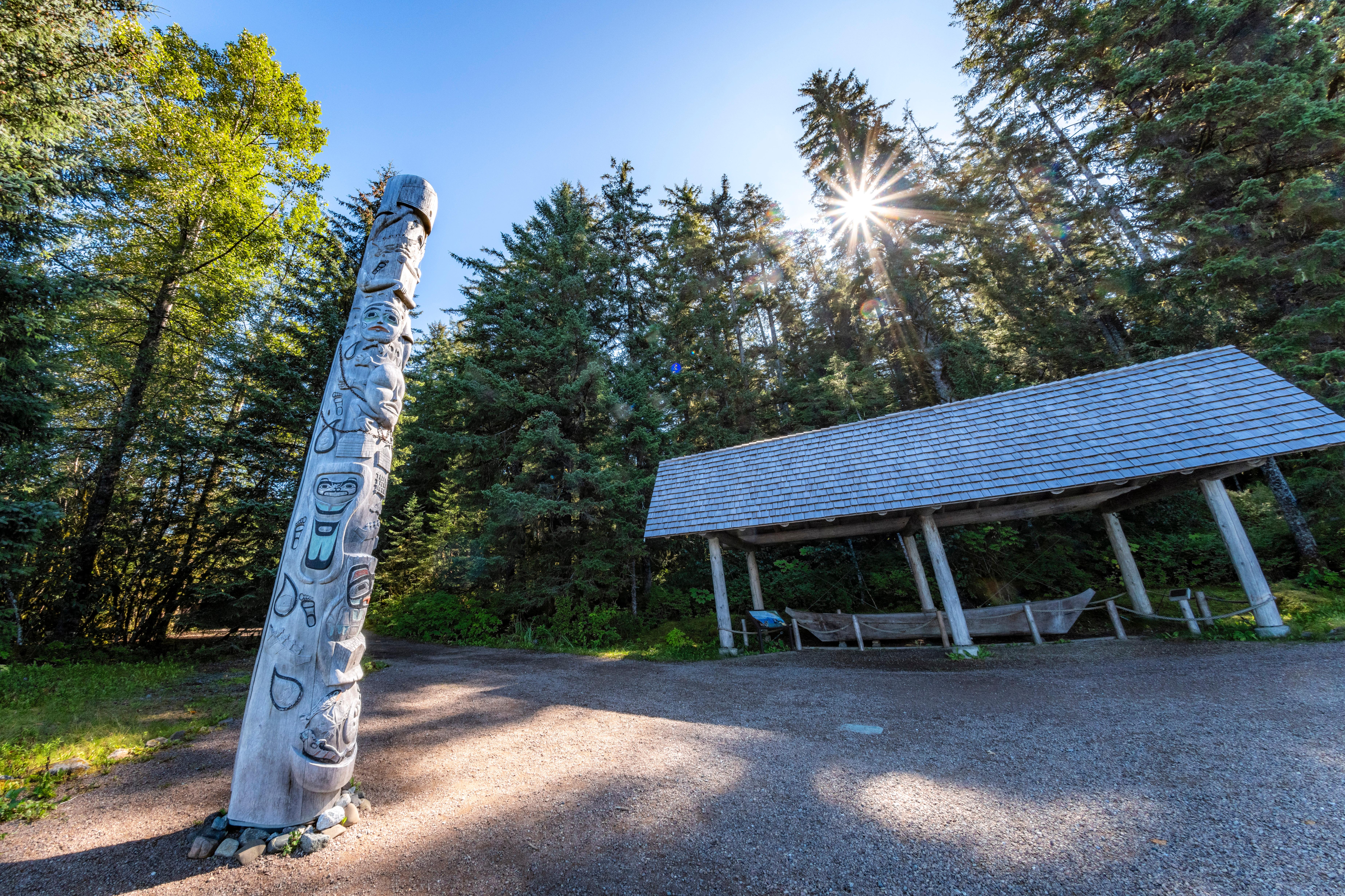 Bright sun shines through trees behind a shelter over a canoe and an intricately carved totem pole.