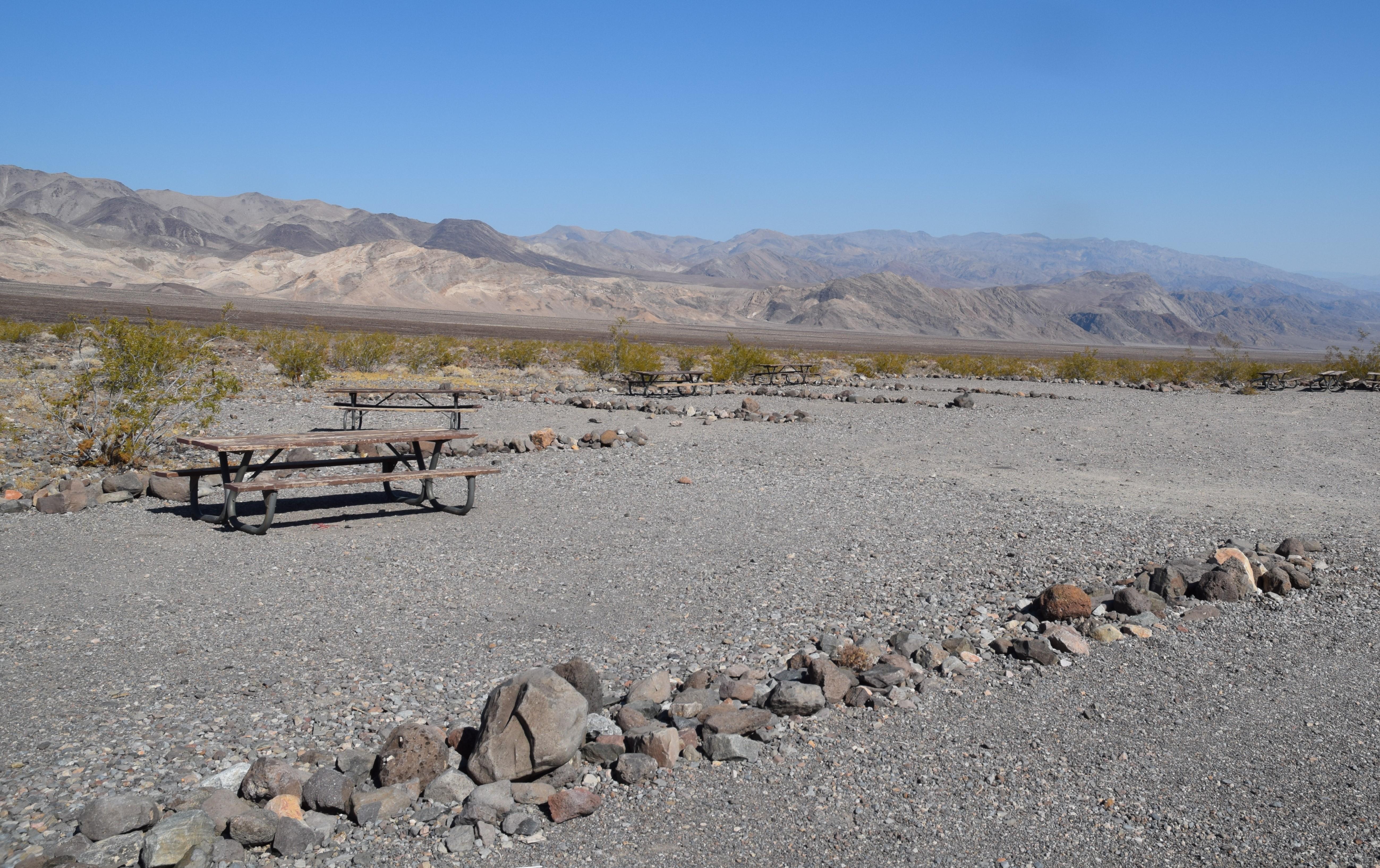 Row of loose 12 inch rocks separate a flat, gravel area. Metal picnic bench on far side.