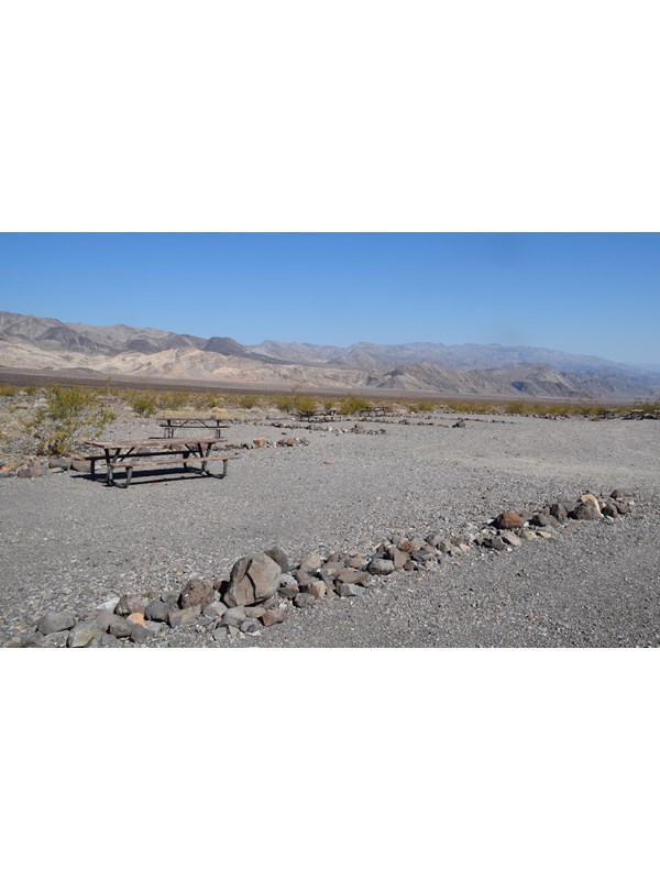 Row of loose 12 inch rocks separate a flat, gravel area. Metal picnic bench on far side.