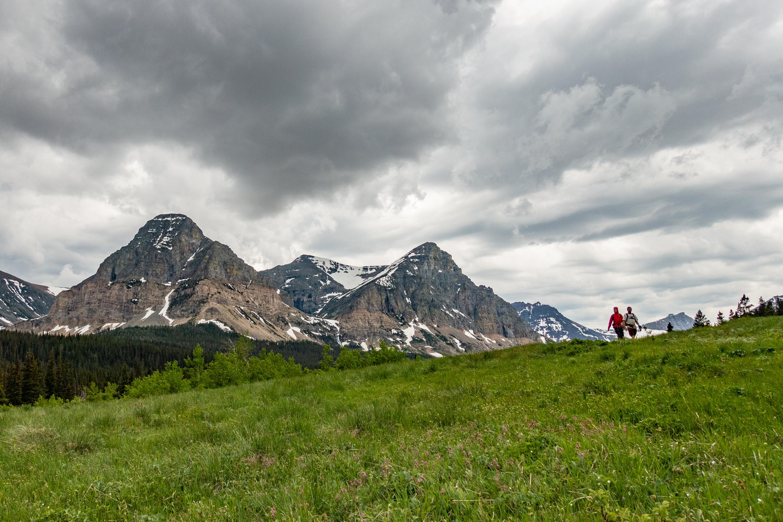 Two people walk down a trail near the Cut Bank Campground under cloudy skies with mountains behind.