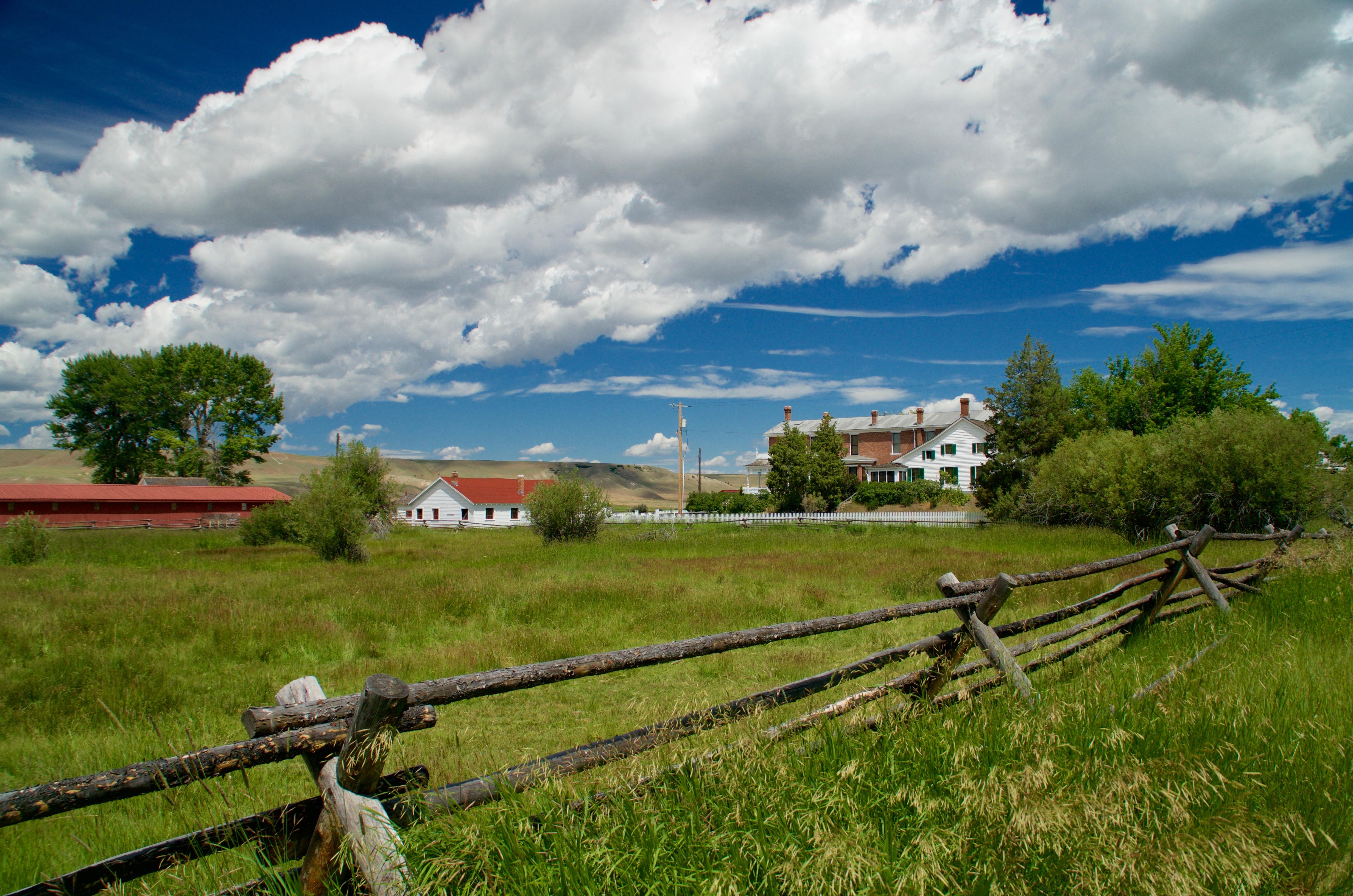 Fenced green pasture in front, 3 ranch buildings across center, blue sky with large white clouds.