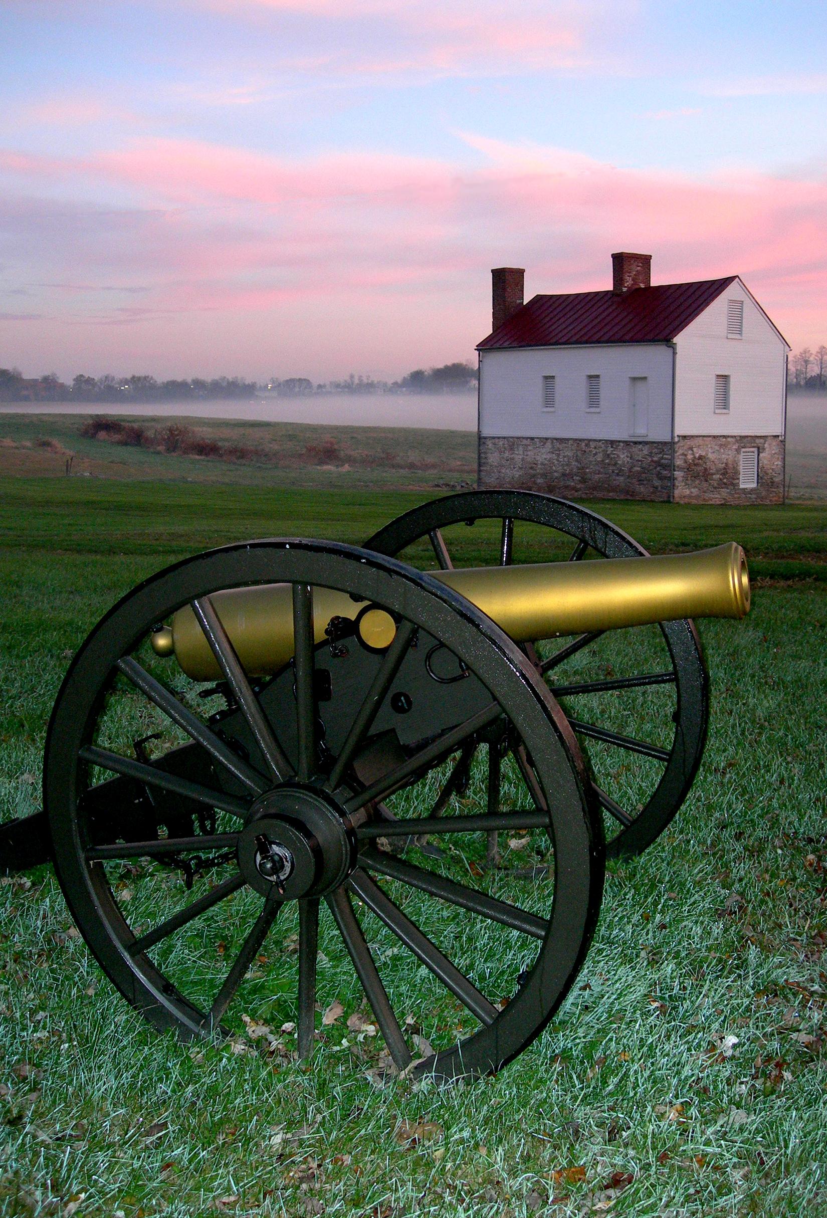A cannon sits in a green field in front of a small white building, the sun is setting.