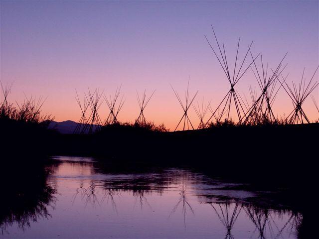 Multiple tepee poles and a river are silhouetted against a dawn sky.