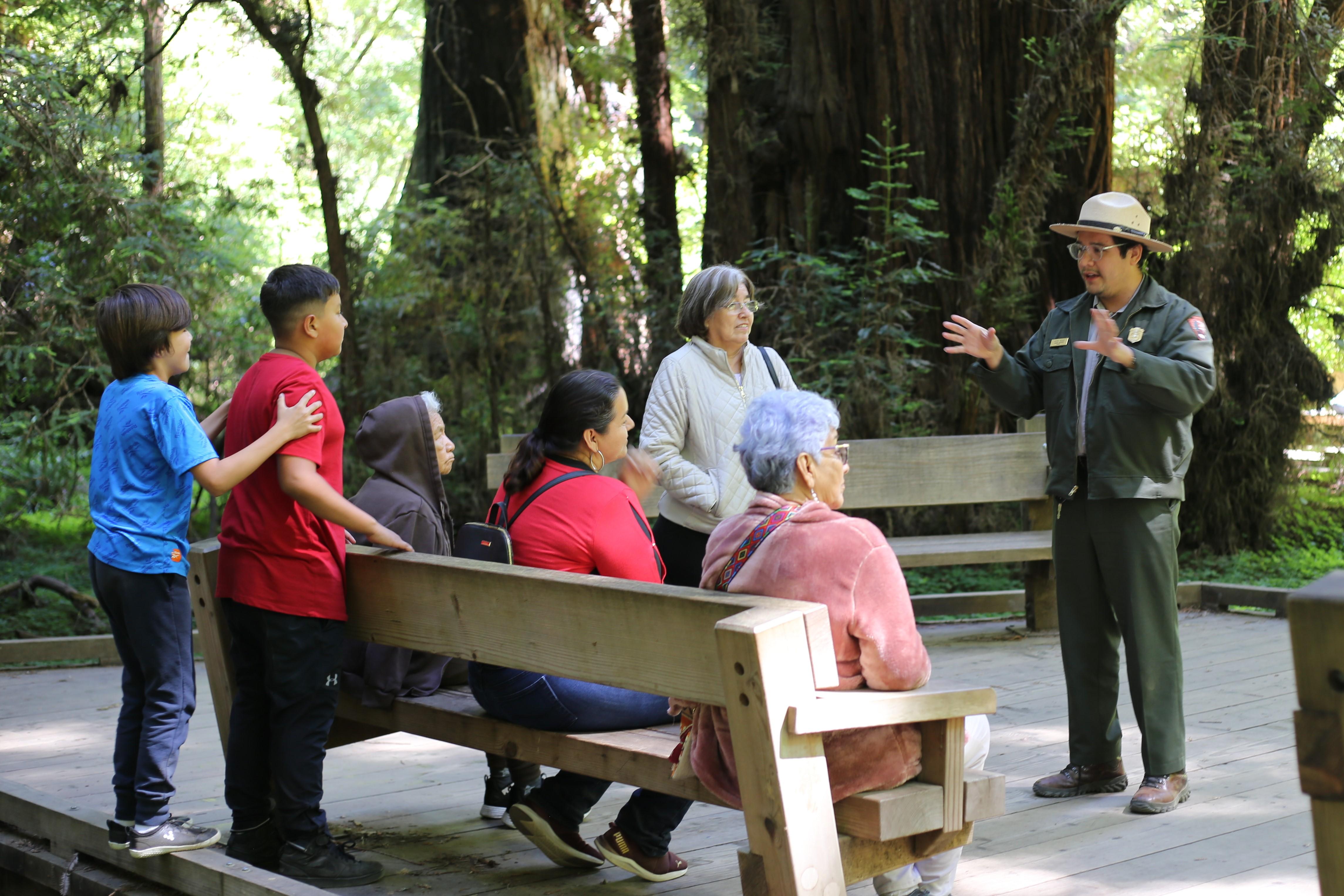 in a shady forest, a ranger in a flat hat and green uniform speaks with a small group of visitors