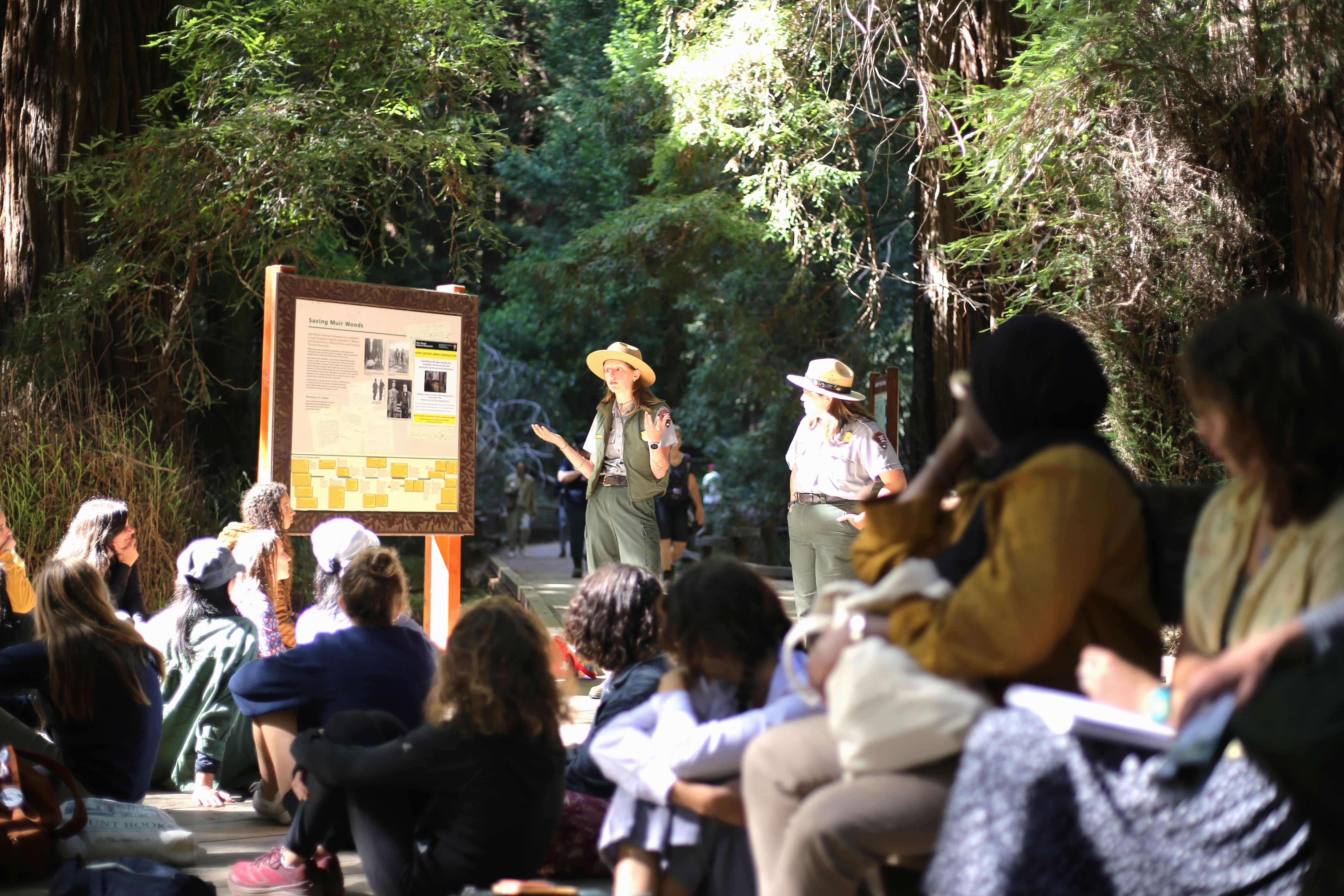 Two rangers stand next to a big sign and speak to visitors seated in the foreground