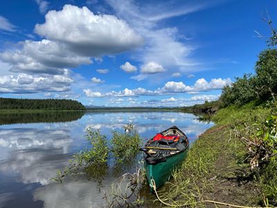 A green pack canoe sits on the bank of the Kobuk River. Clouds are mirrored on the water\'s surface.
