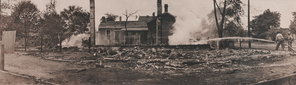 Historic photo of two people using a hose to extinguish a burning building. 