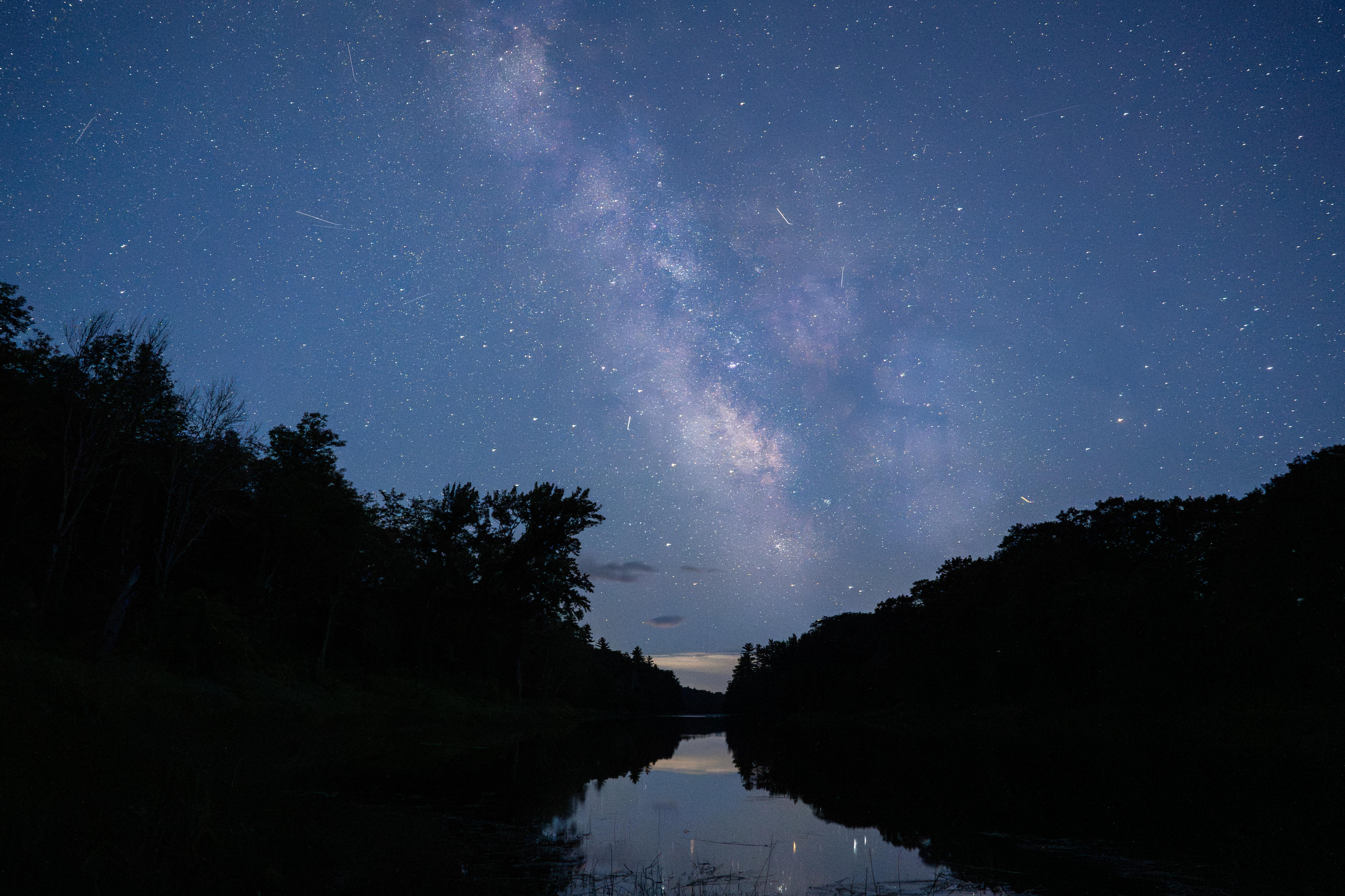 A blue night sky filled with stars sits over reflective water. Dark trees line the image.