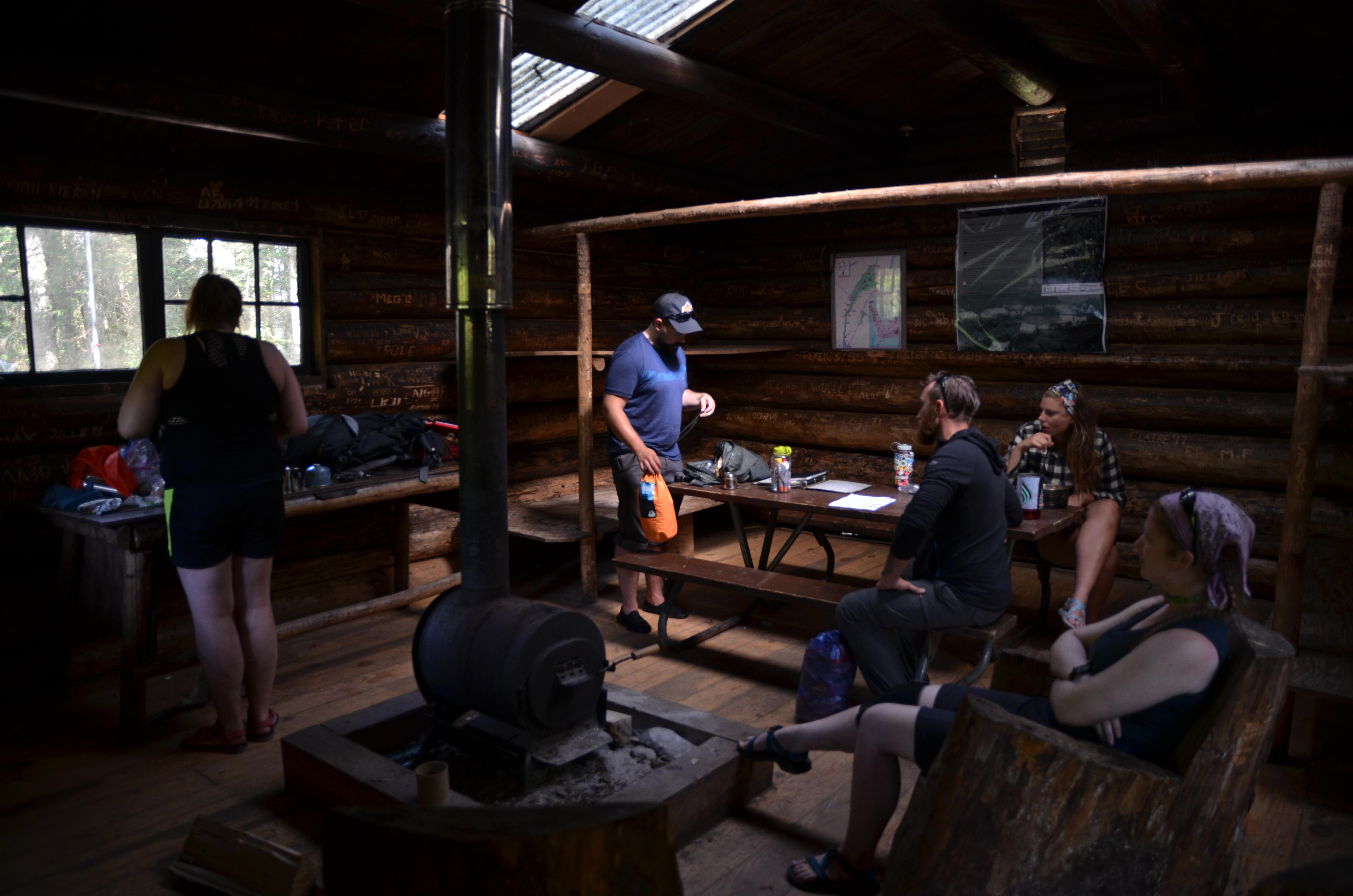 Inside of a log cabin with people gathered around a wood stove.