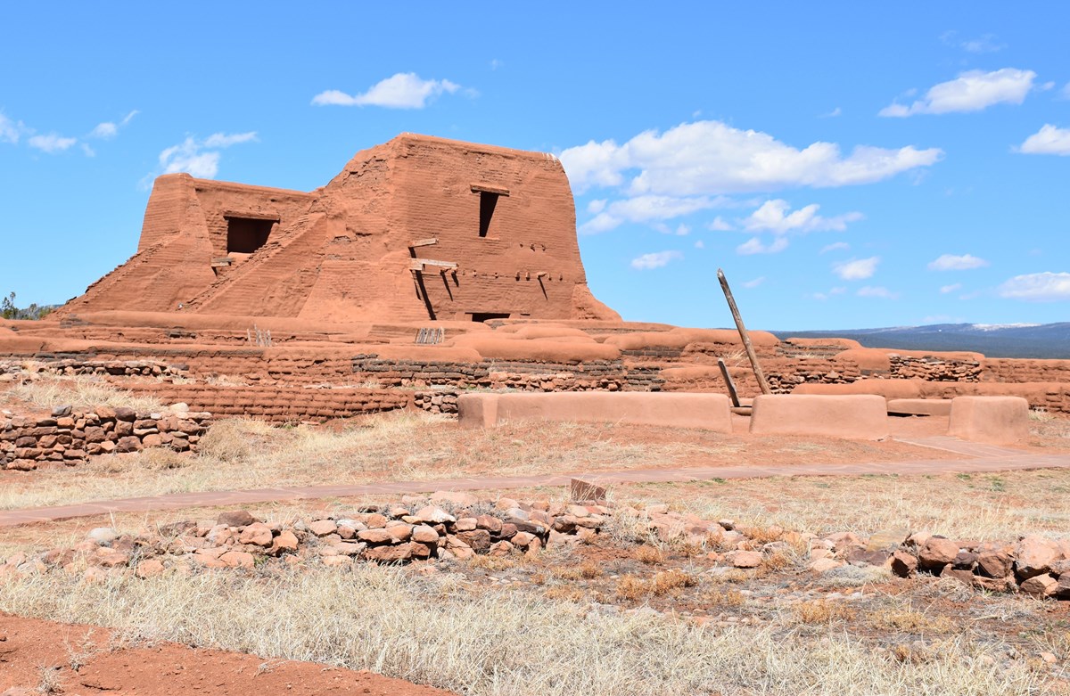 Large brown adobe mission church with low brick walls and kiva ladder in front