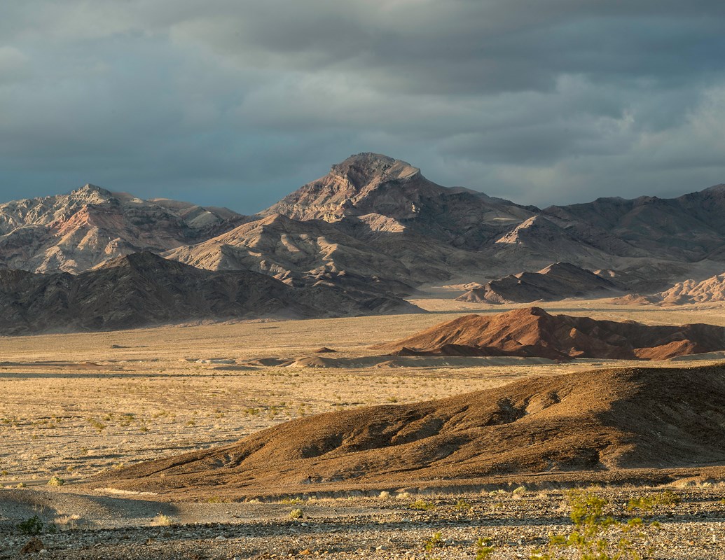Rugged gray mountains rise over a brown desert landscape under a gray sky.