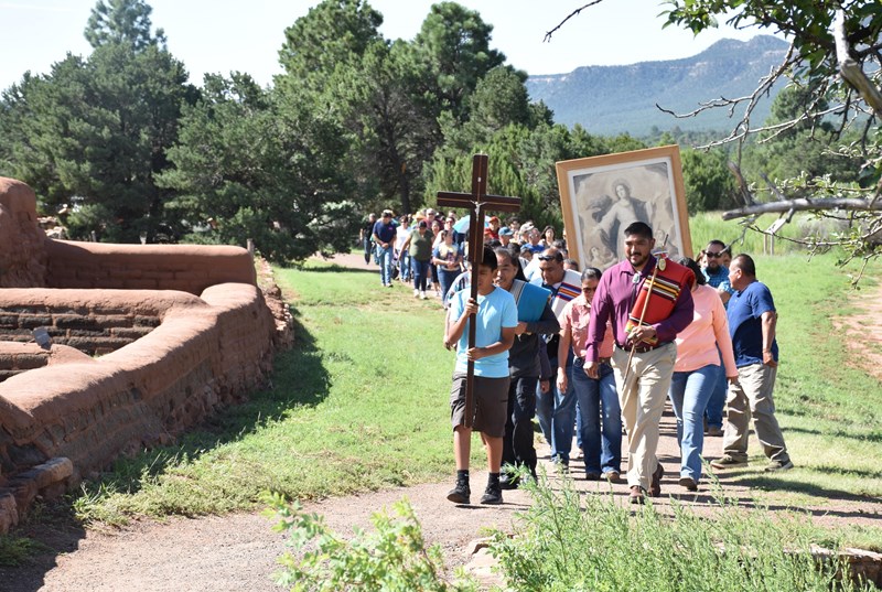 Feast Day Procession A line of people walk past adobe bricks and trees on a path