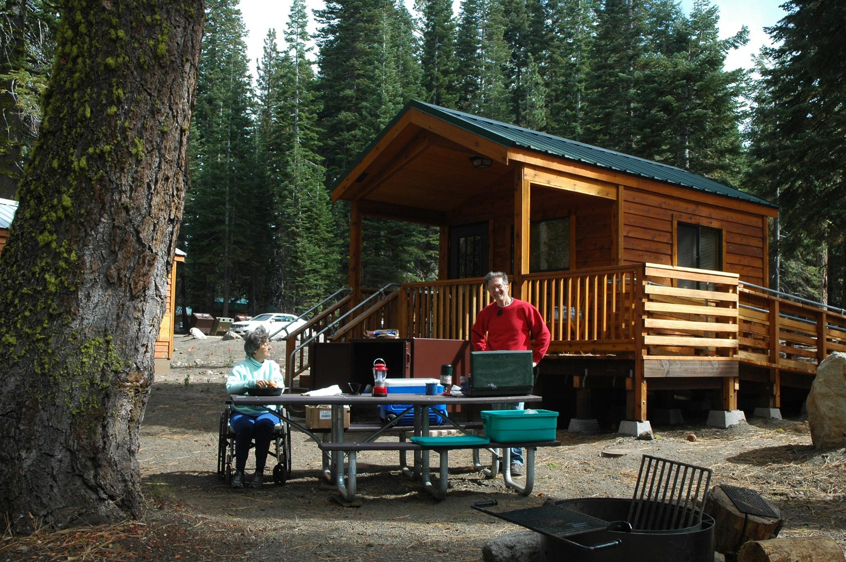 A woman sits in a wheelchair and a man stands at a picnic table backed by a small, wooden cabin.