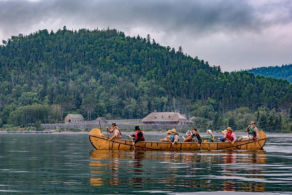 People in historic clothing paddle a canoe in a bay in front of a stockade.