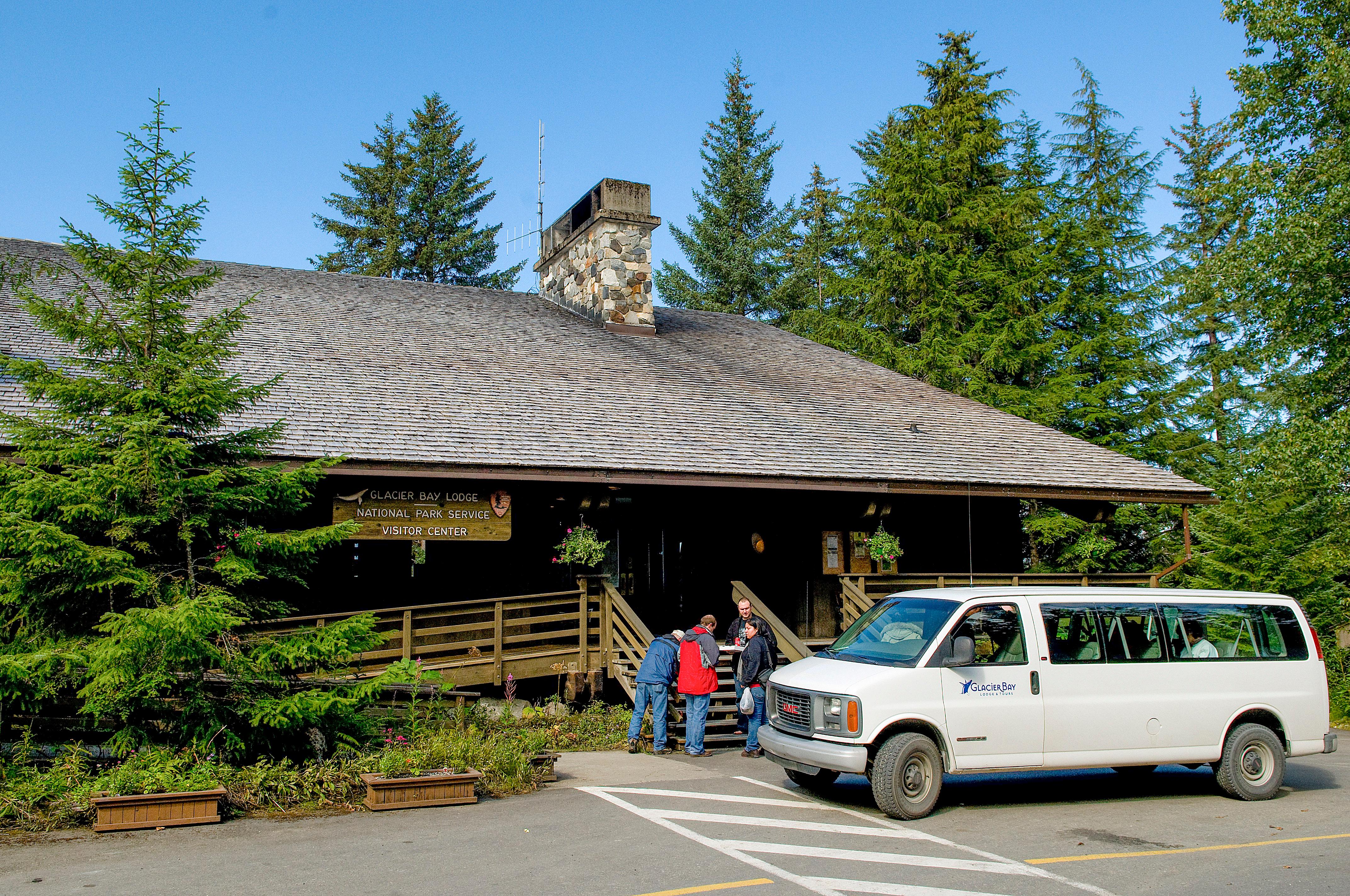 A glacier bay lodge van sits in front of the Glacier Bay Lodge entrance, several visitors stand by.