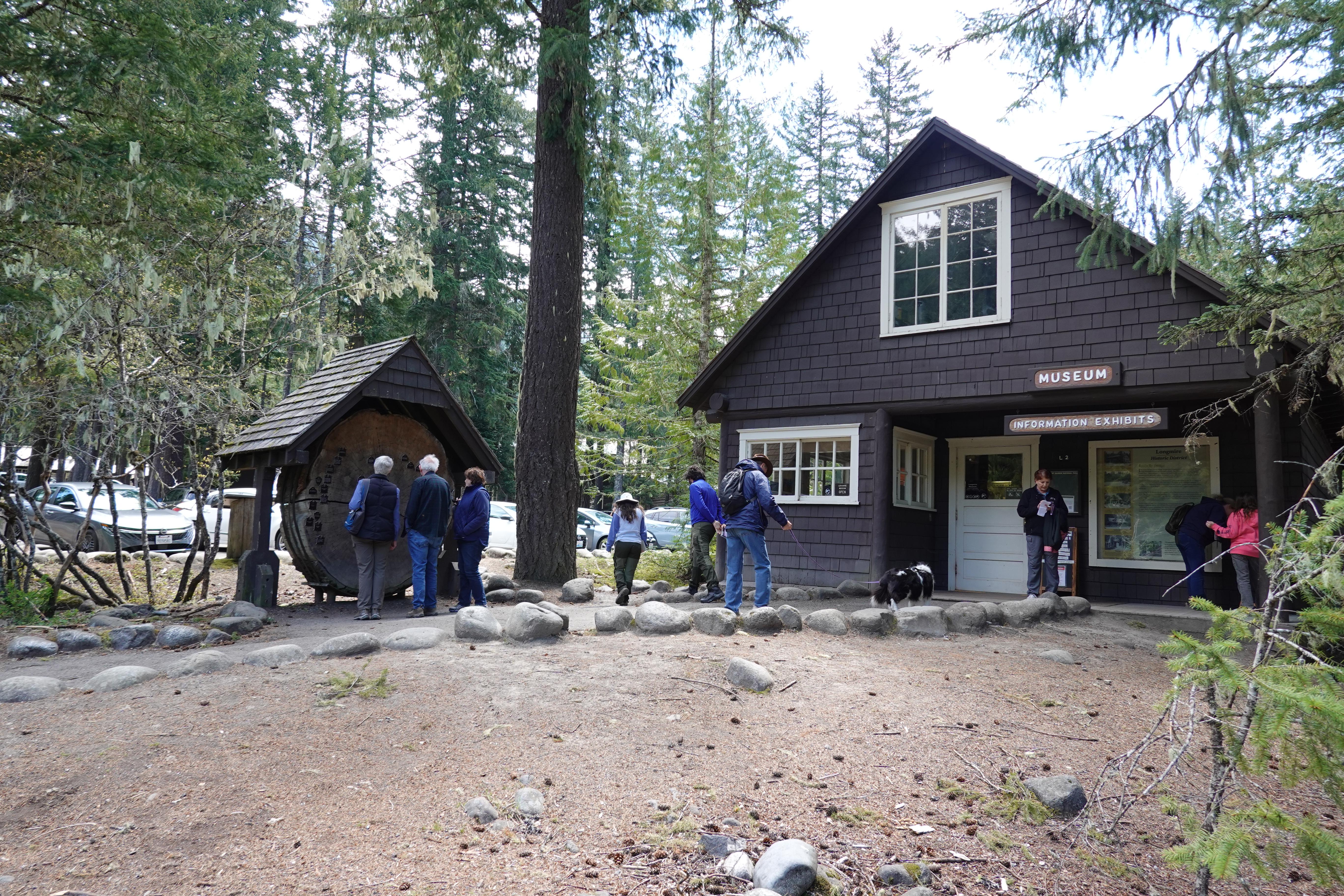 Visitors outside of a small wood building look at signs and a cross section of a large log.