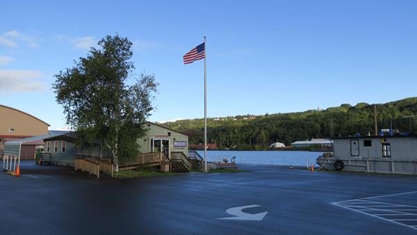 A building is surrounded by a parking lot, tree, and flag pole nestled next to a large body of water