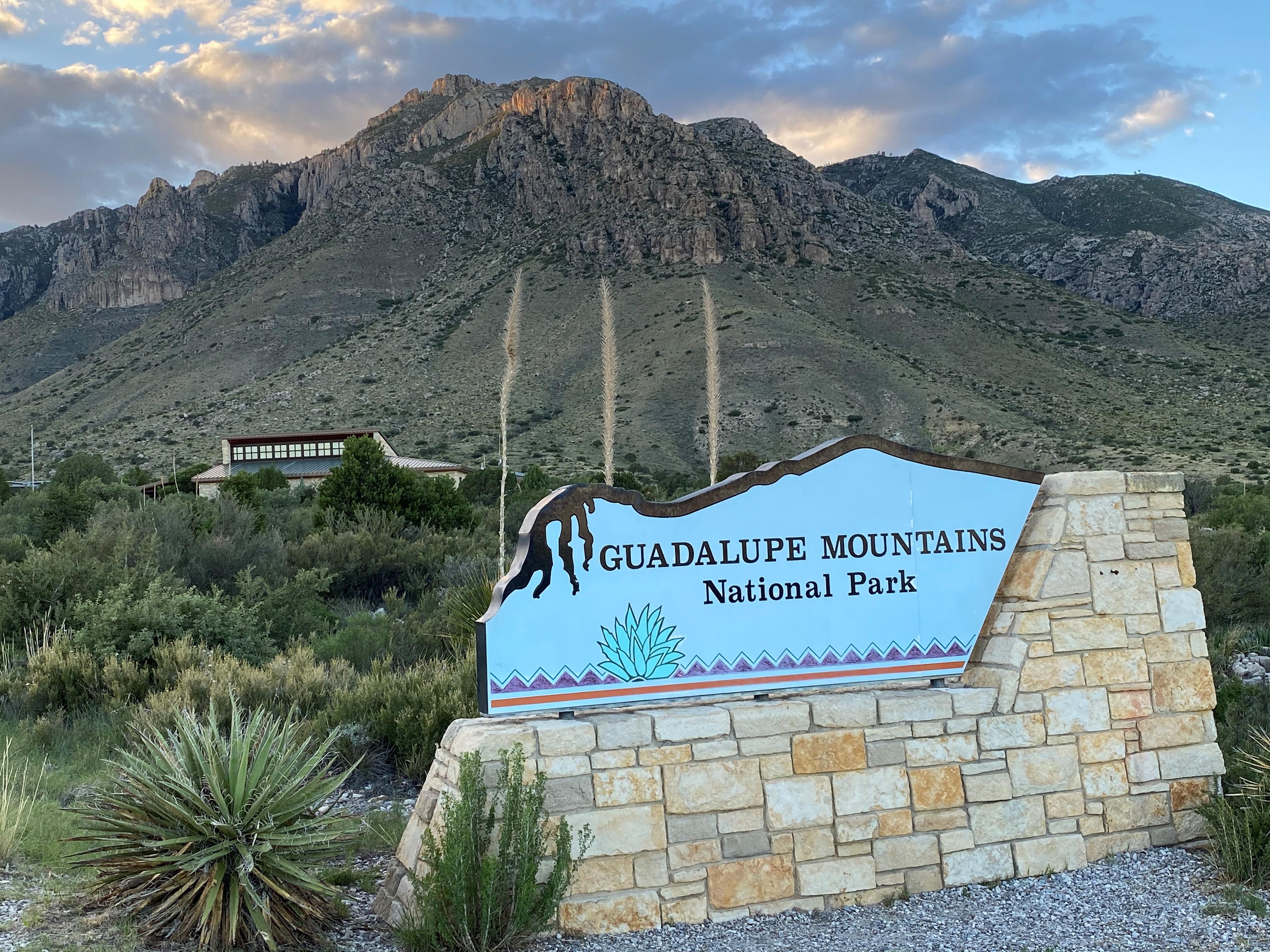 A metal and stone entrance sign stands in front of a building and desert mountains