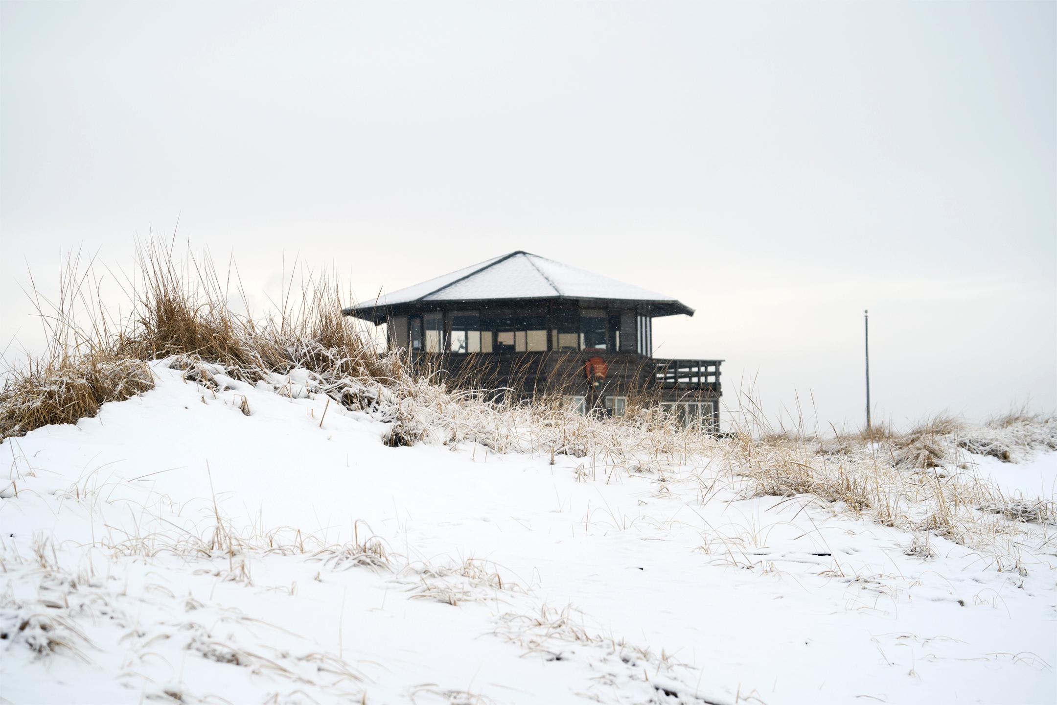 A brown hexagonal building covered in snow stands behind a beachside dune.