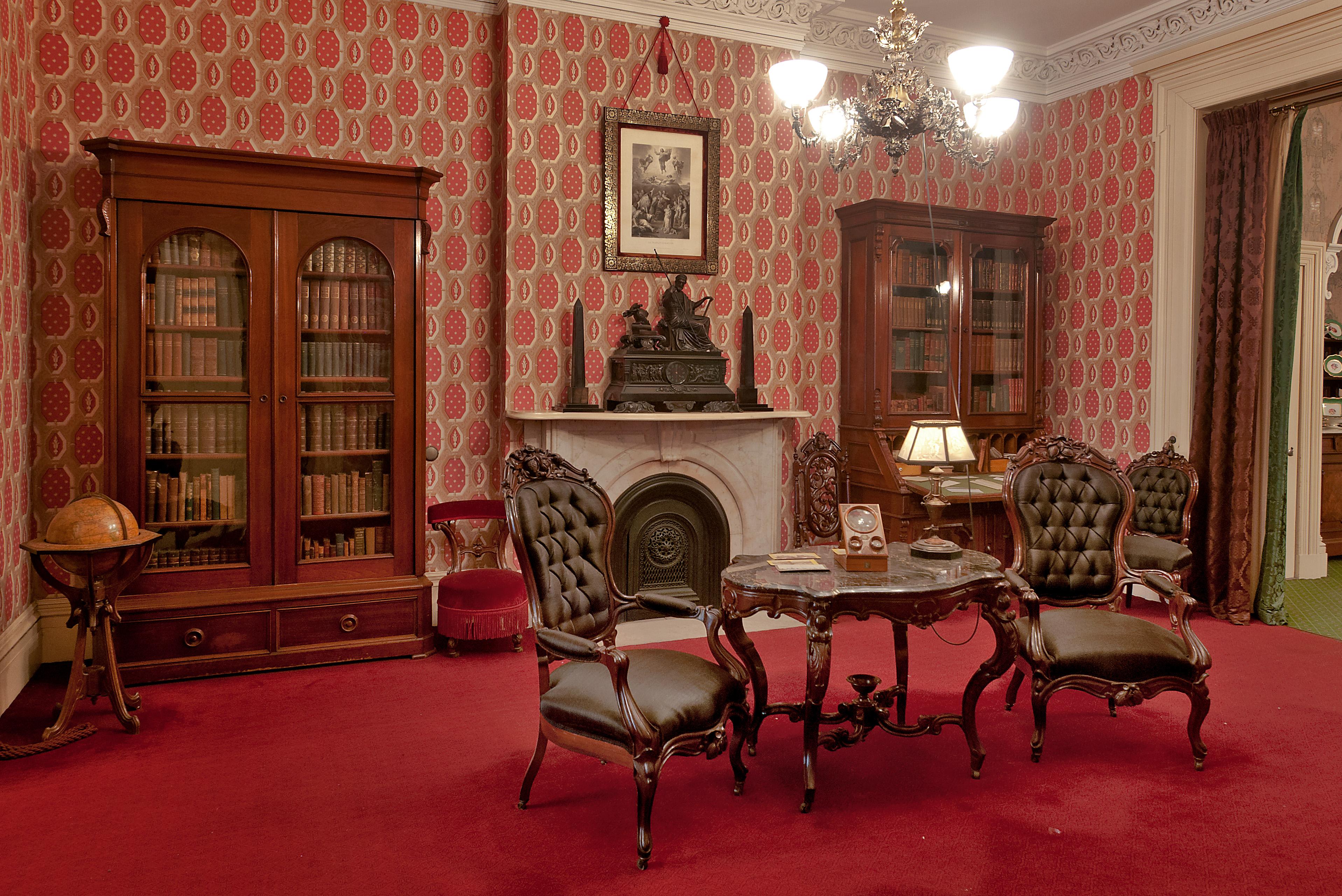 A room with red wallpaper, red carpeting, and wooden Victorian-era furniture.