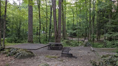 A tent pad, picnic table, and fire ring near rocks, trees, and other sites in the distance.