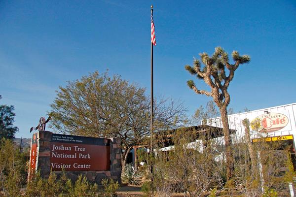 A sign that reads, "Joshua Tree National Park Visitor Center", a flag pole, vegetation, and building