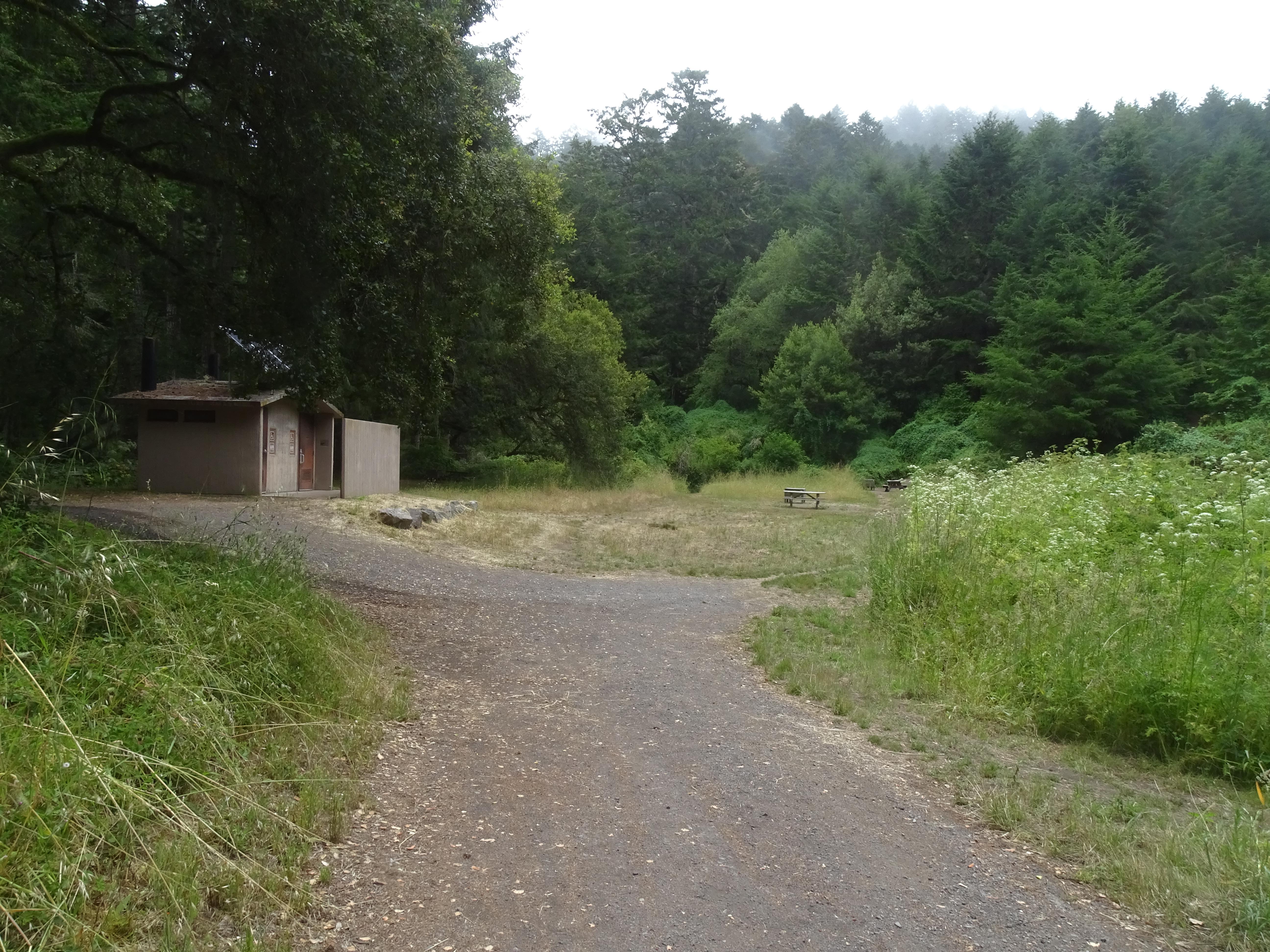 In a wooded valley, a leads into a campground with a vault toilet building under trees on the left.