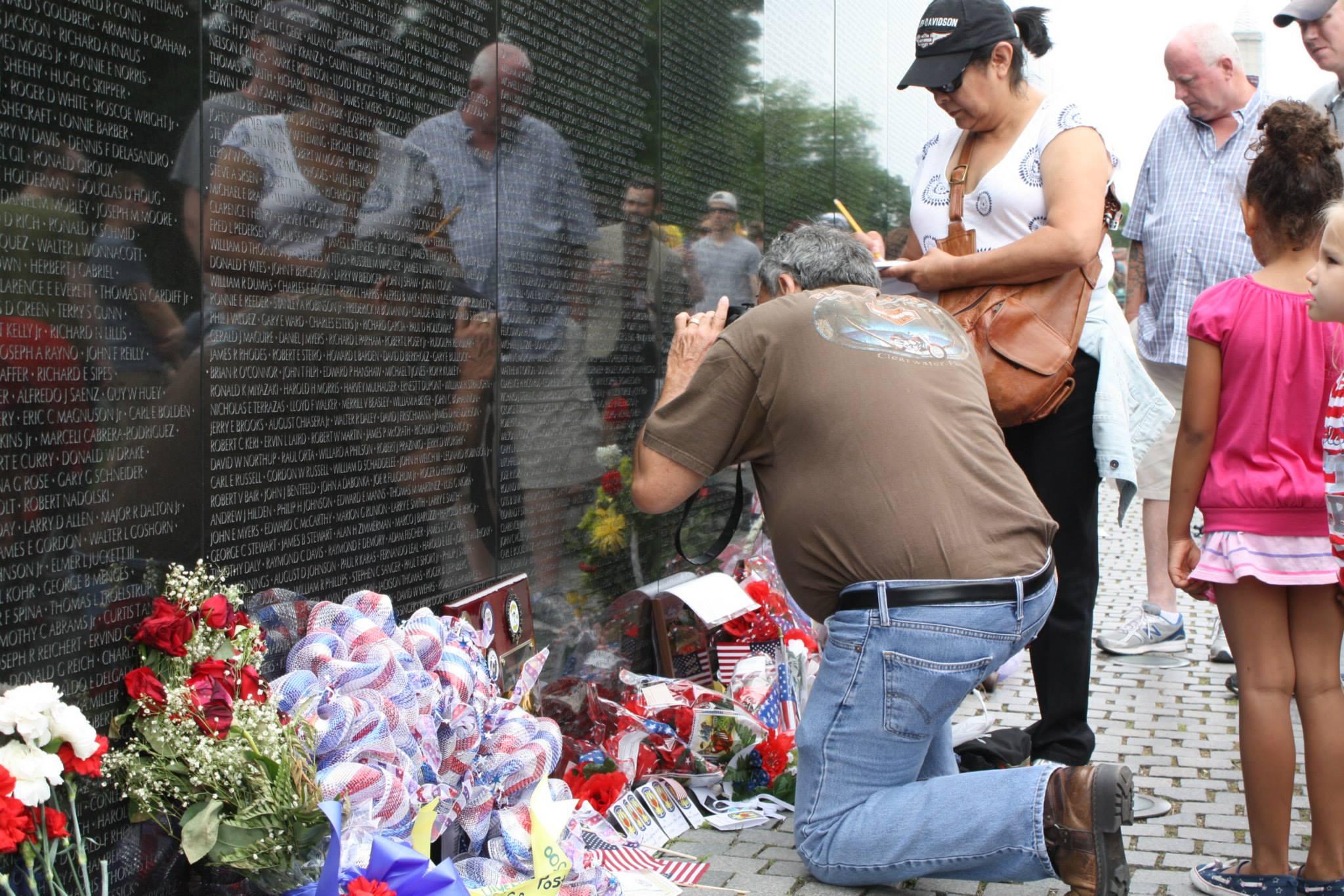A man kneels and takes a photo of the wall surrounded by other people. Flowers are at the wall base.