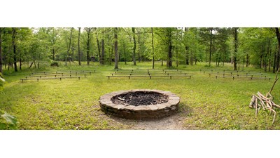 A large stone campfire circle is surrounded by a semicircle of benches in the woods.