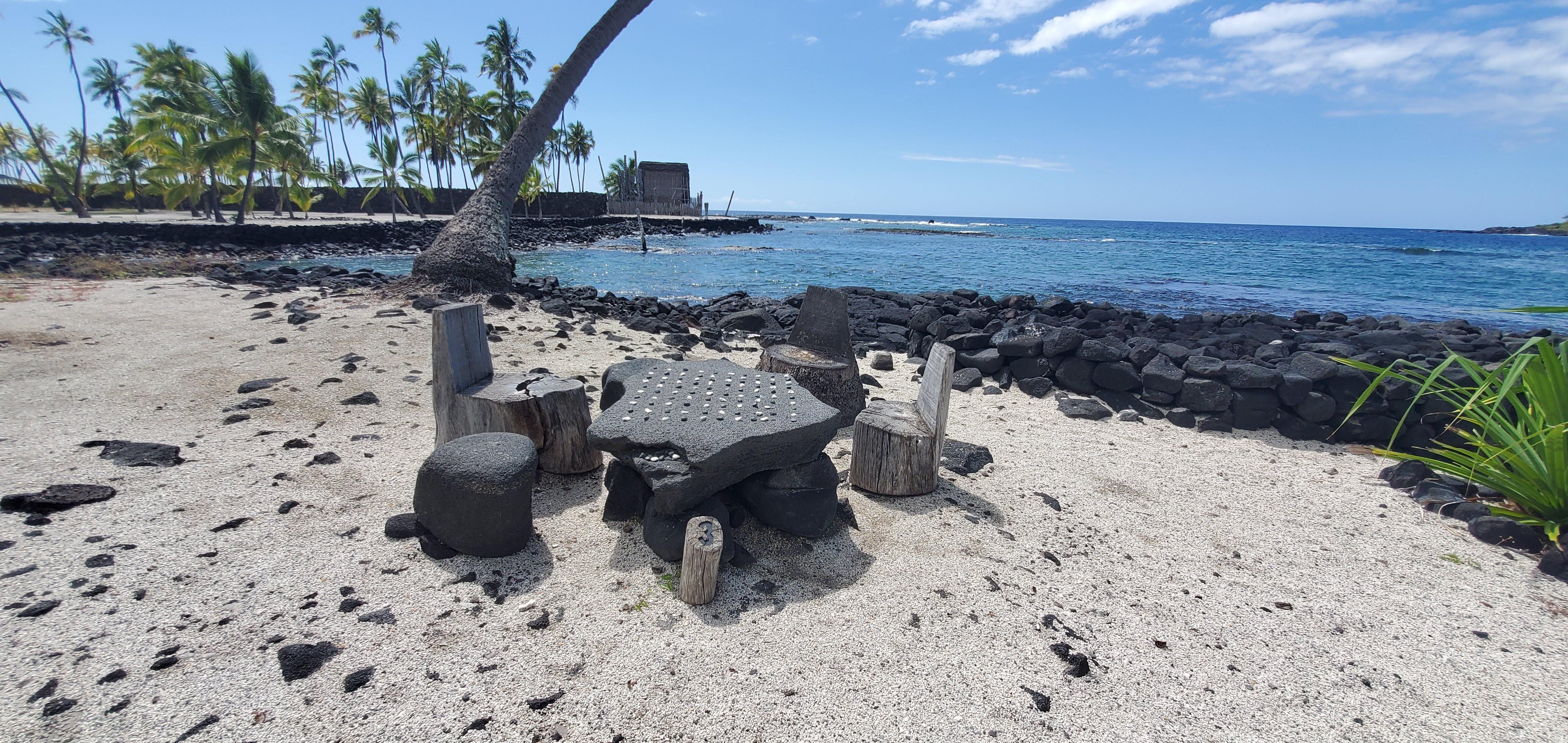 A stone papamū (Kōnane playing surface) with Hale o Keawe and Hōnaunau Bay in the background.