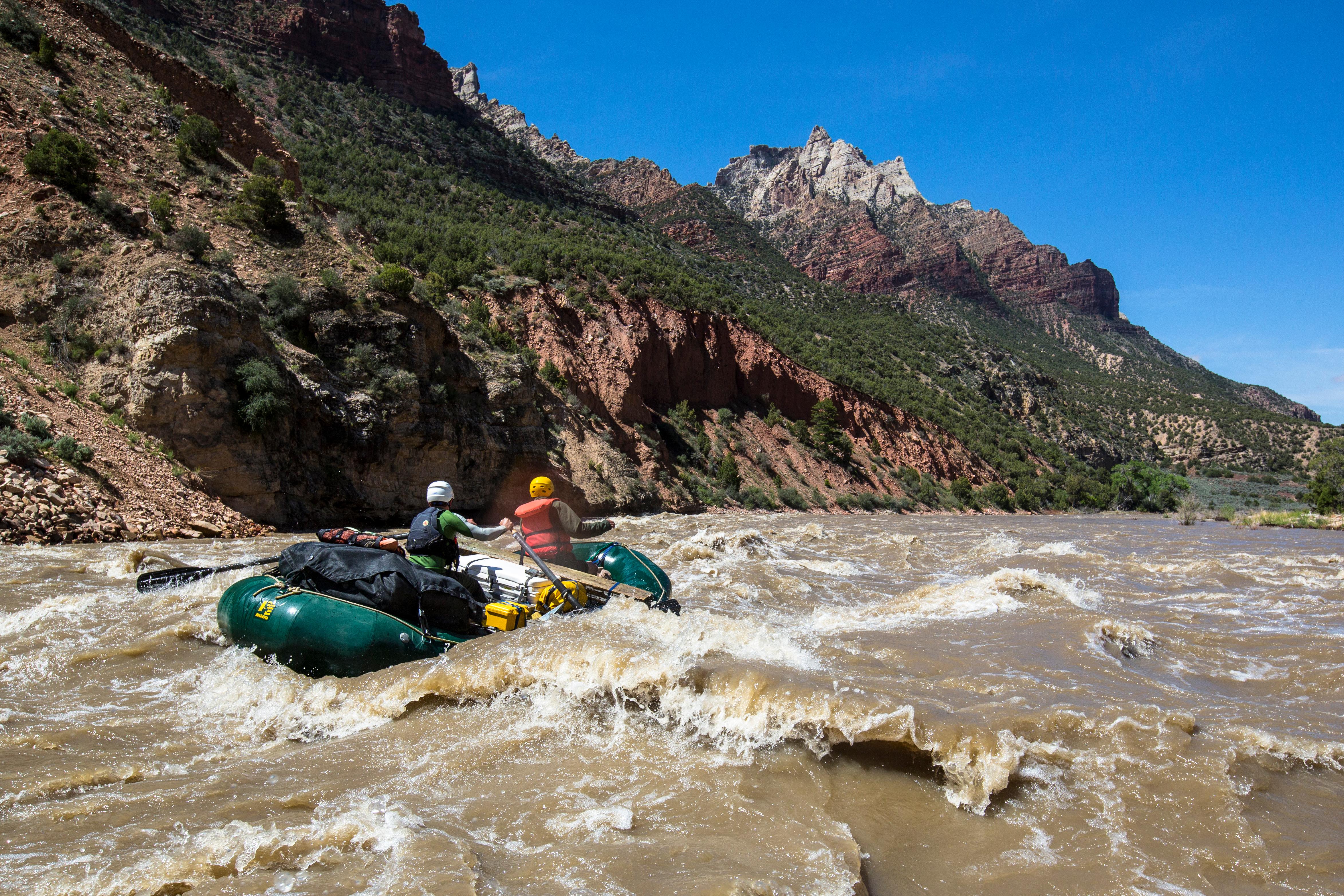 A green rubber raft floats over a rapid on a brown colored river in front of multi-colored mountains