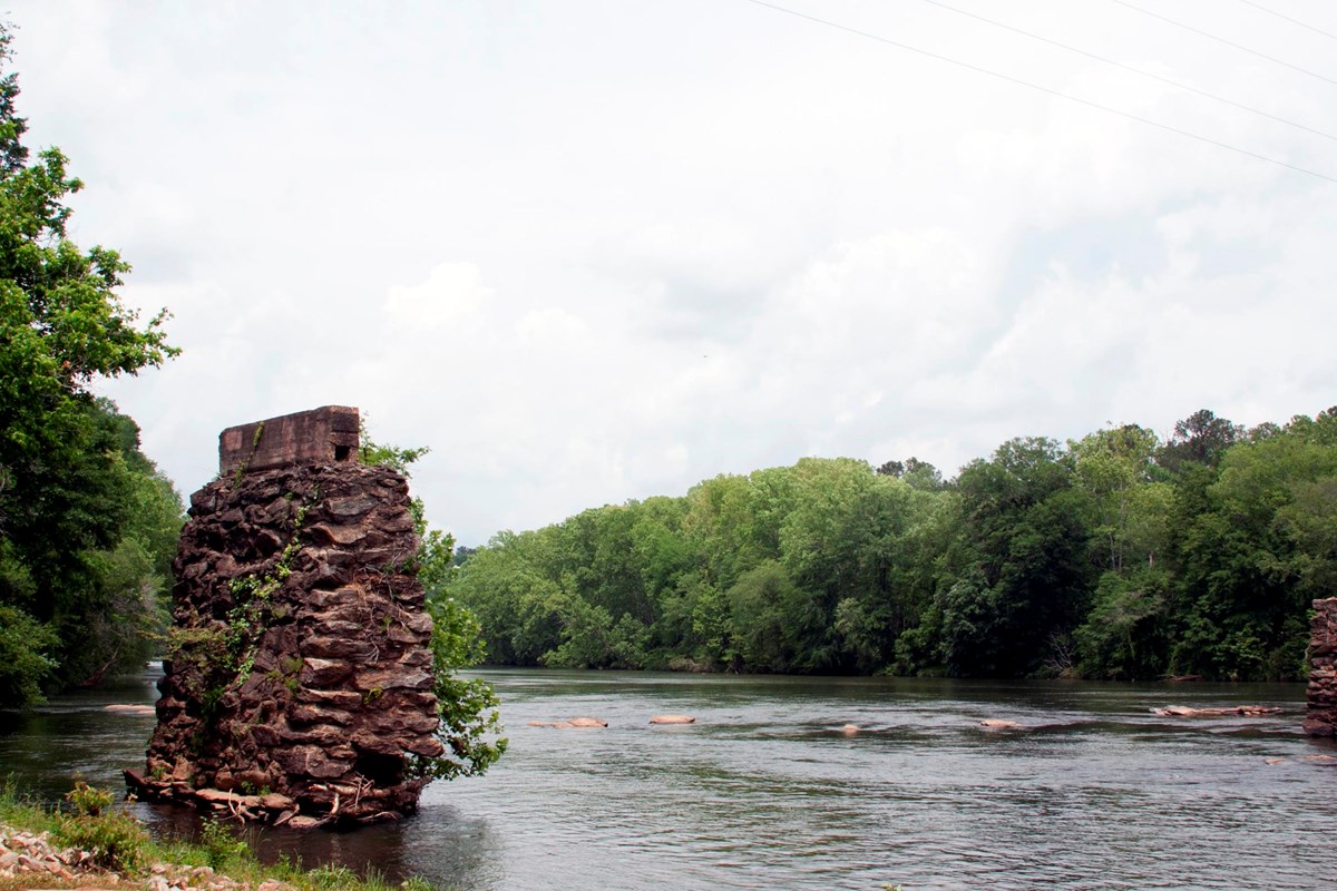 flat river, green trees lining banks, old bridge pier made of stones in river on left side of photo