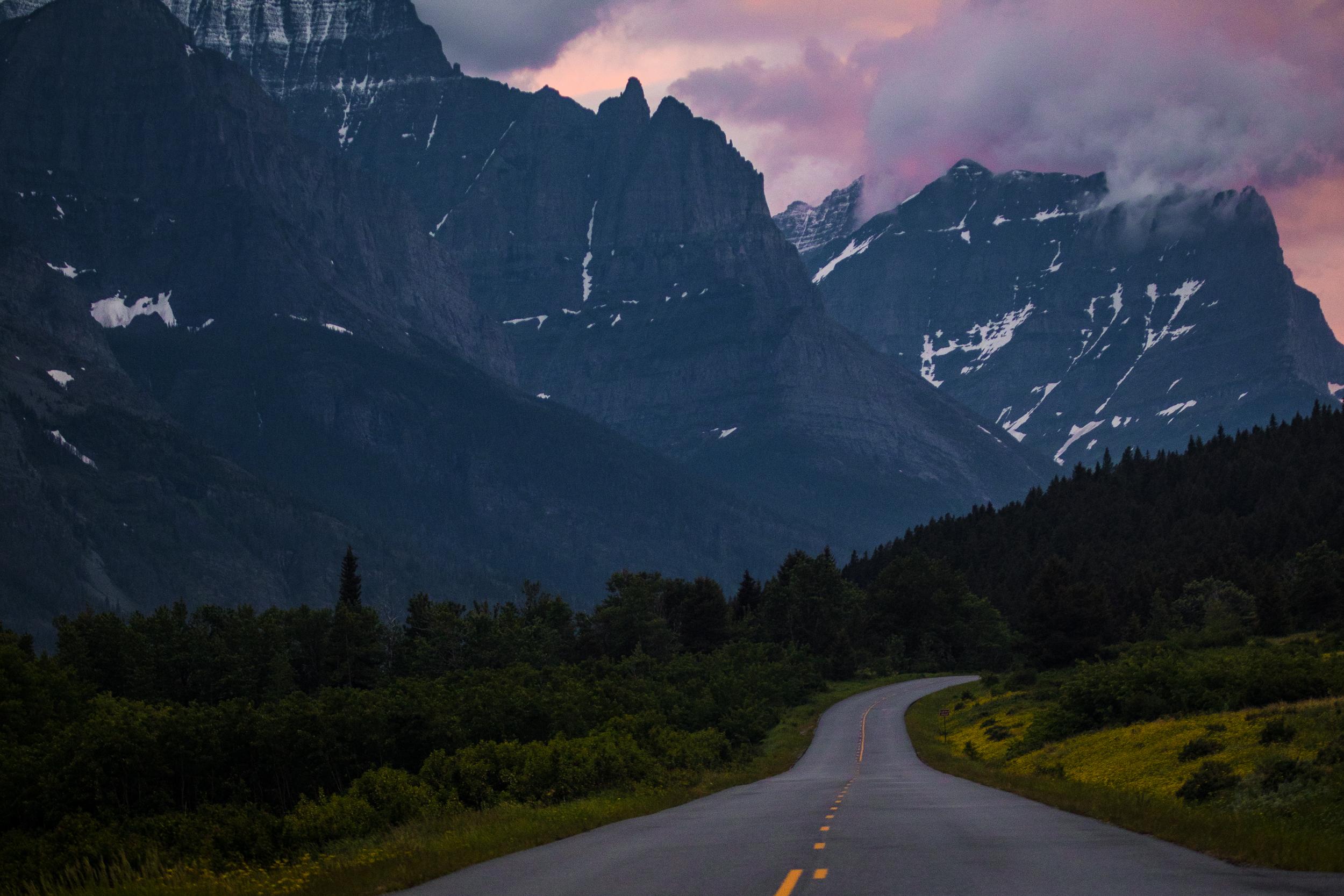 Jagged peaks rise out of a forested valley and an empty road curves off into the distance.