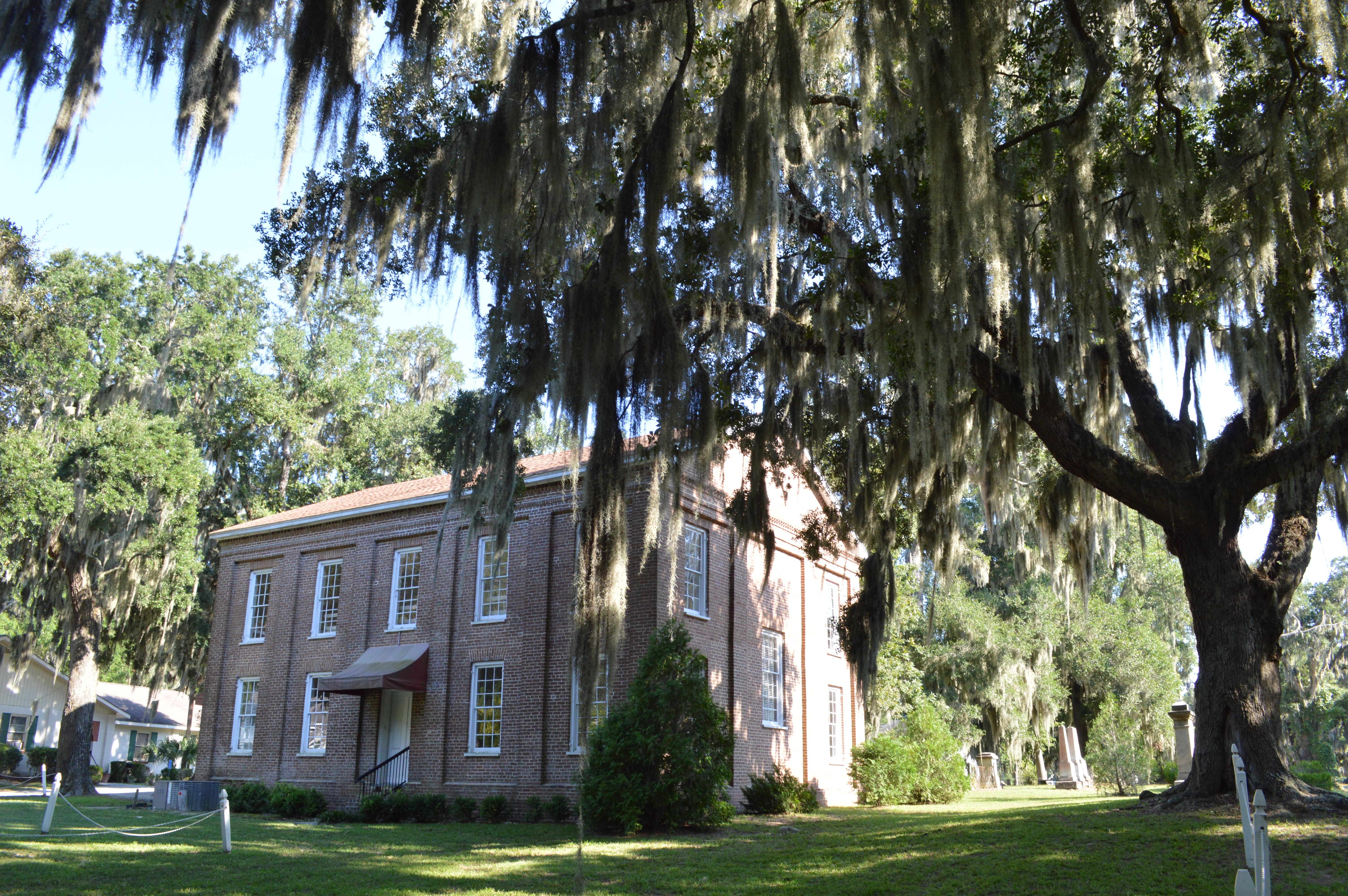 A two story brick building flanked by large live oak trees