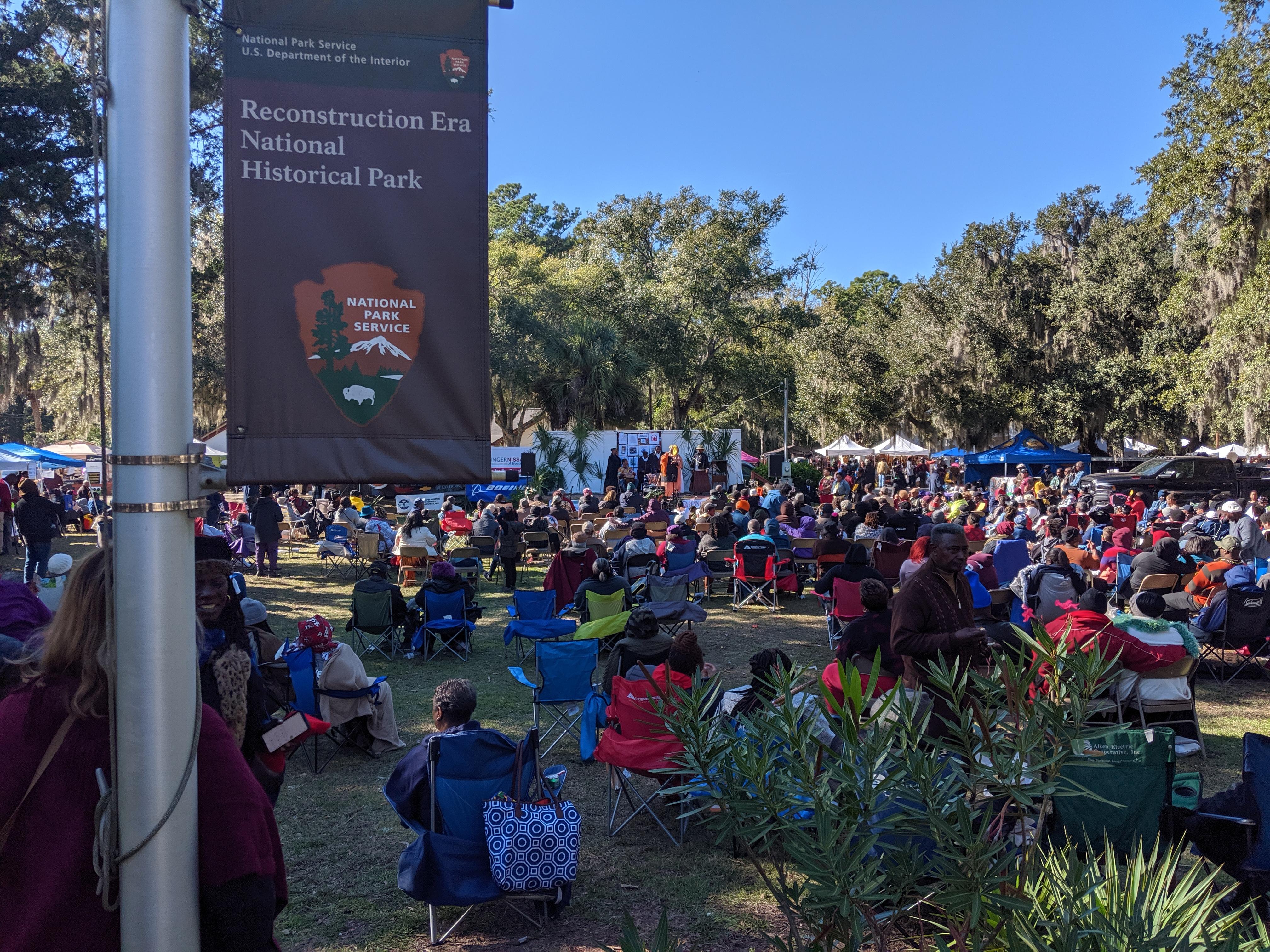 A large crowd of people gather in a field. In the foreground is an NPS sign