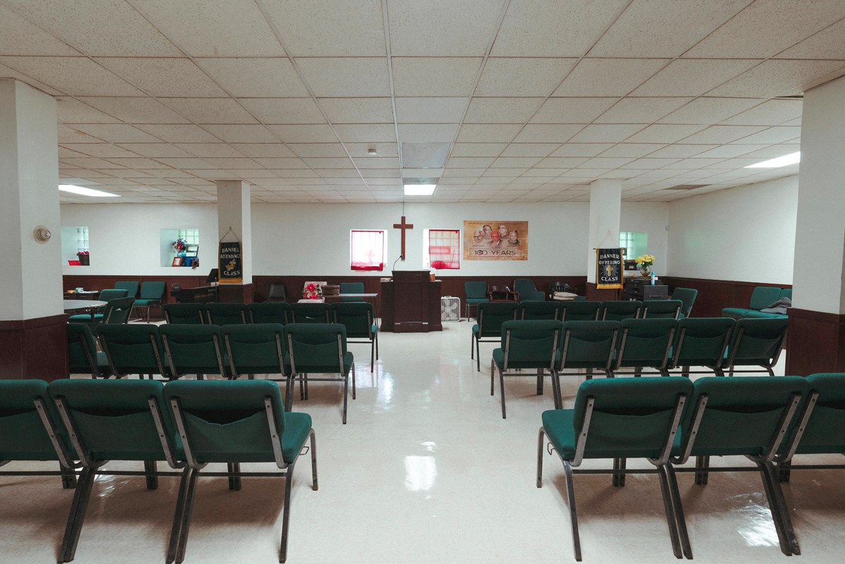 Rows of green seats in a large white room pointed toward a pulpit and a cross on a wall. 