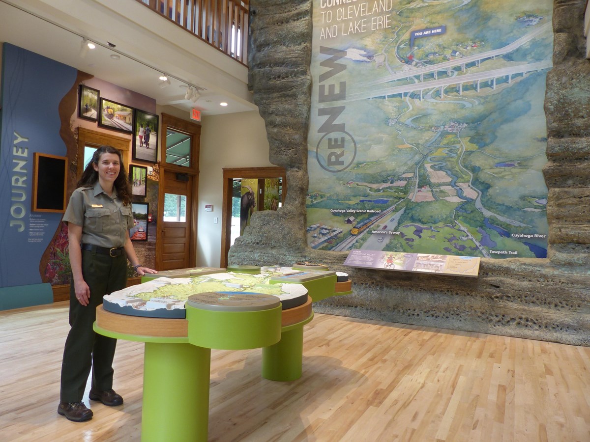 A park ranger stands beside a terrain model of the Cuyahoga Valley with a mural behind.