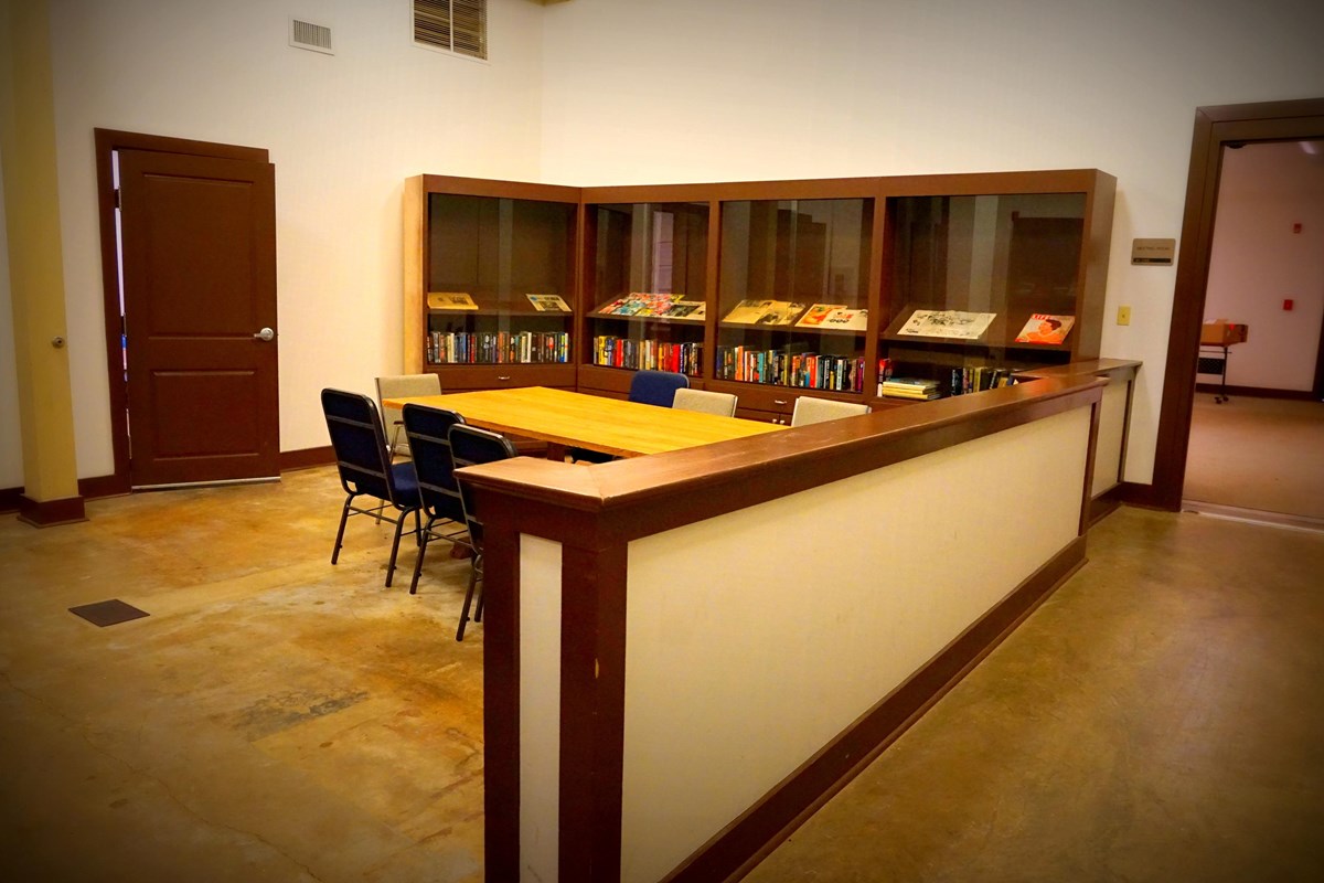 A table sits besides shelves featuring magazines and books in a white room. 