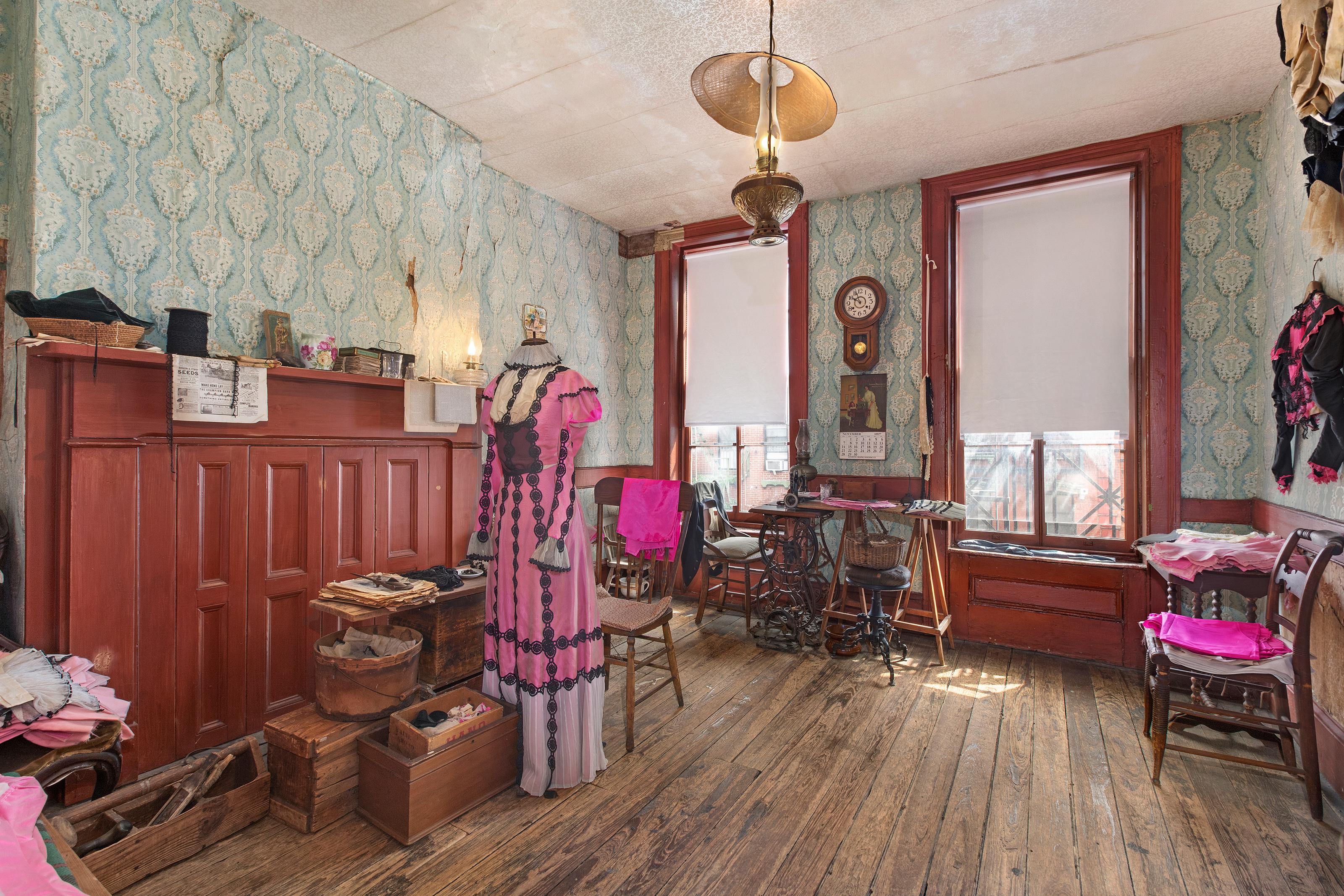 A room with wooden floors, blue wall paper, two windows, some furniture and a dress hanging.