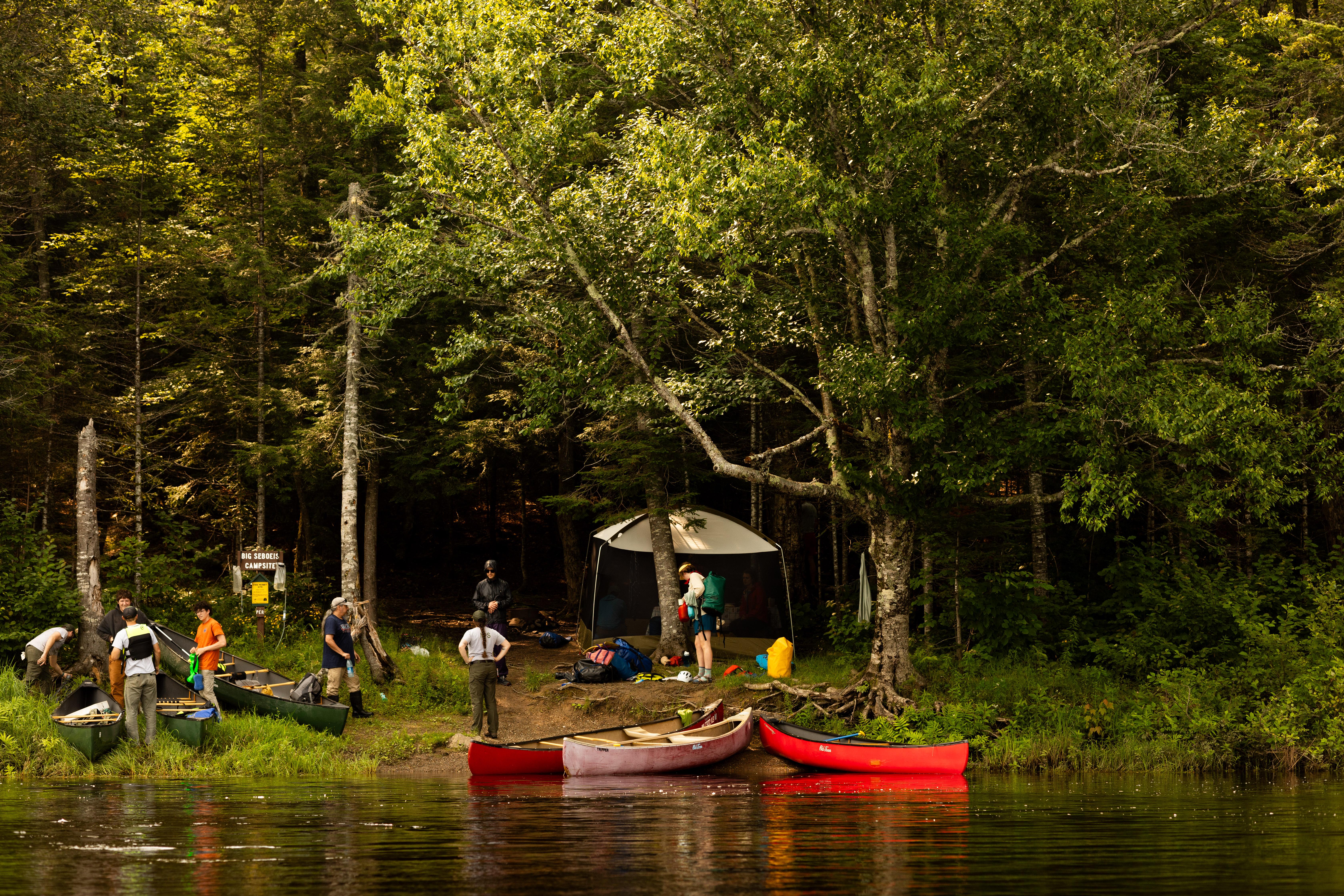 A group of campers with their canoes along the shore of a river. A shaded forest is behind them.