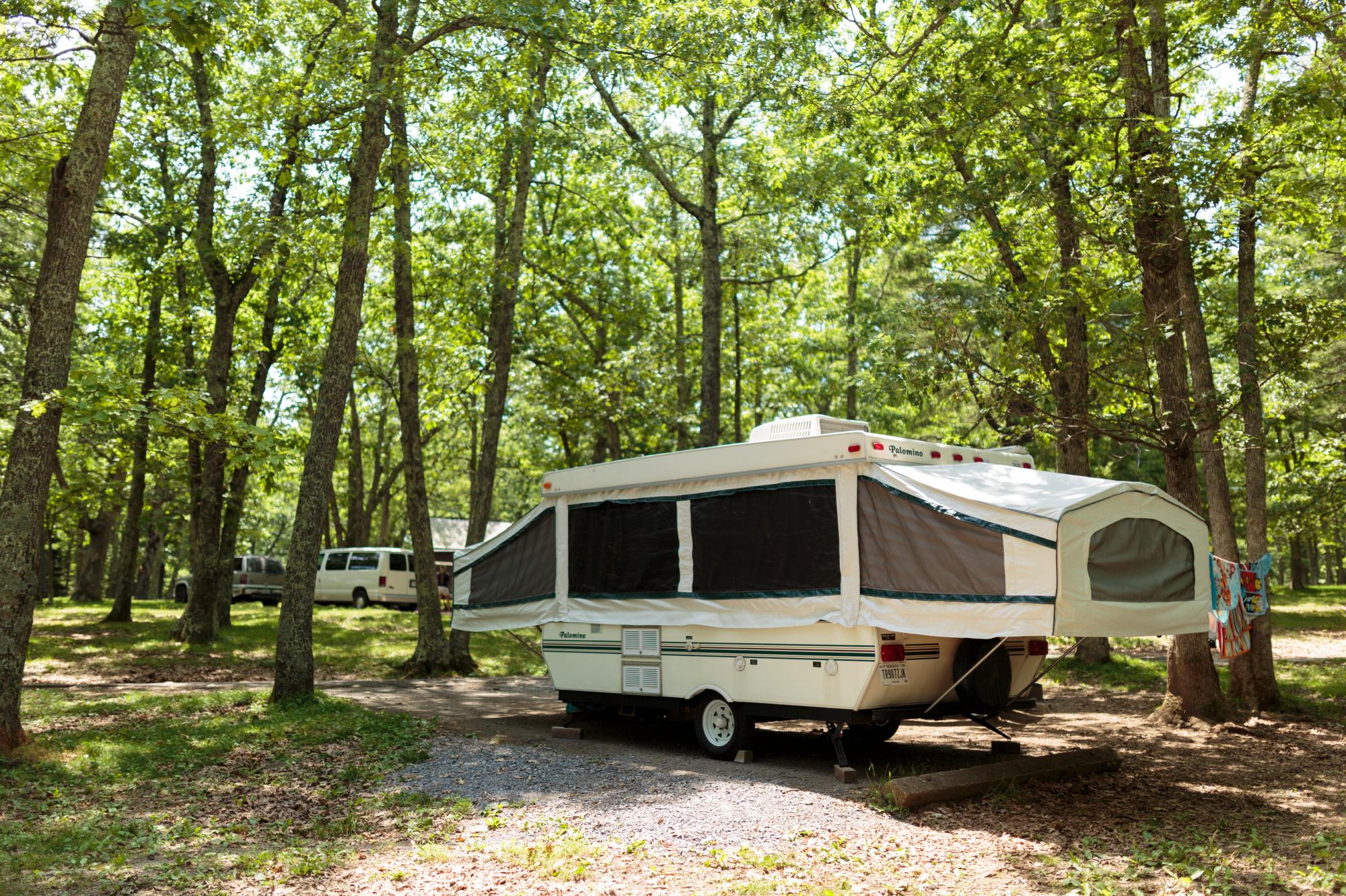 A small pop-up camper is parked in a campground with green trees overhead.