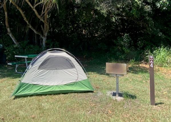 A green tent is pitched next to a raised grill on a grassy area. Trees cast some shade from behind.