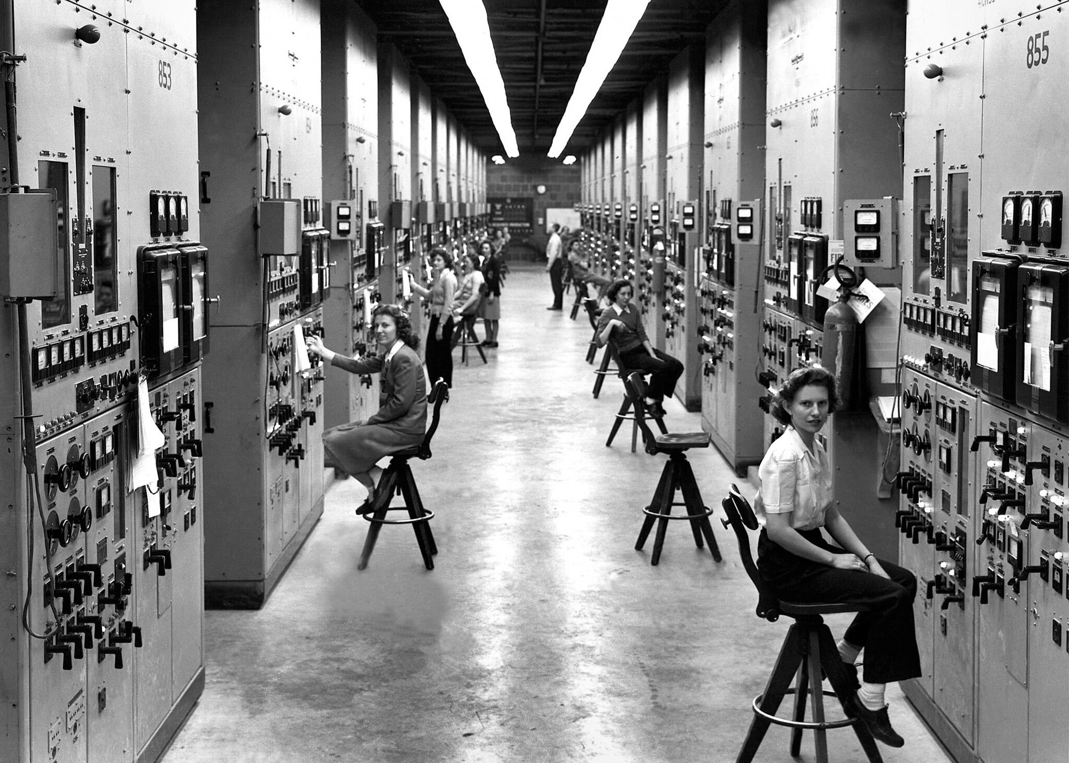 Several women sit on stools along a long hallway full of dials and meters.